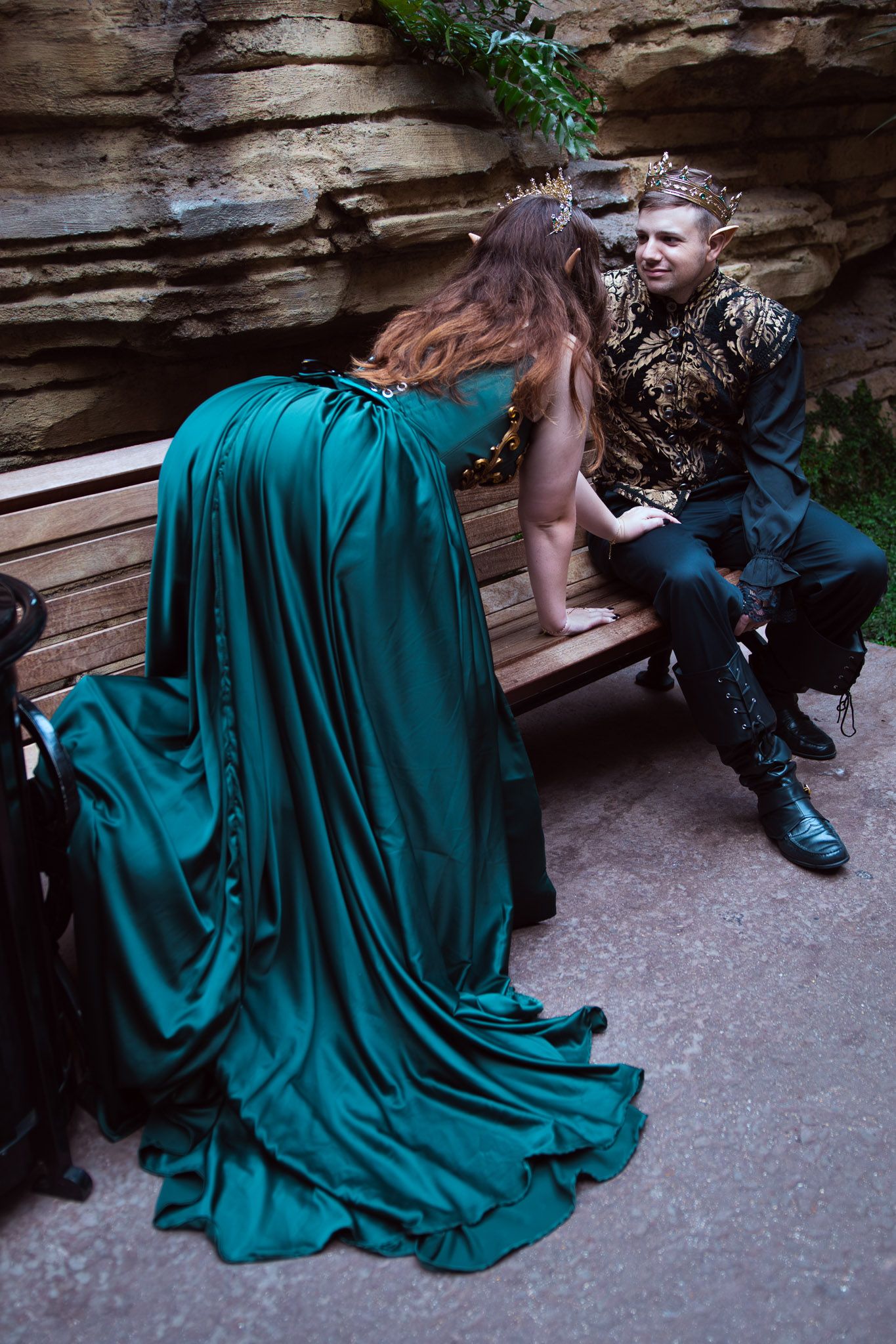 A woman in a flowing emerald green gown crawls before a man wearing a gold crown on a bench near stone walls.
