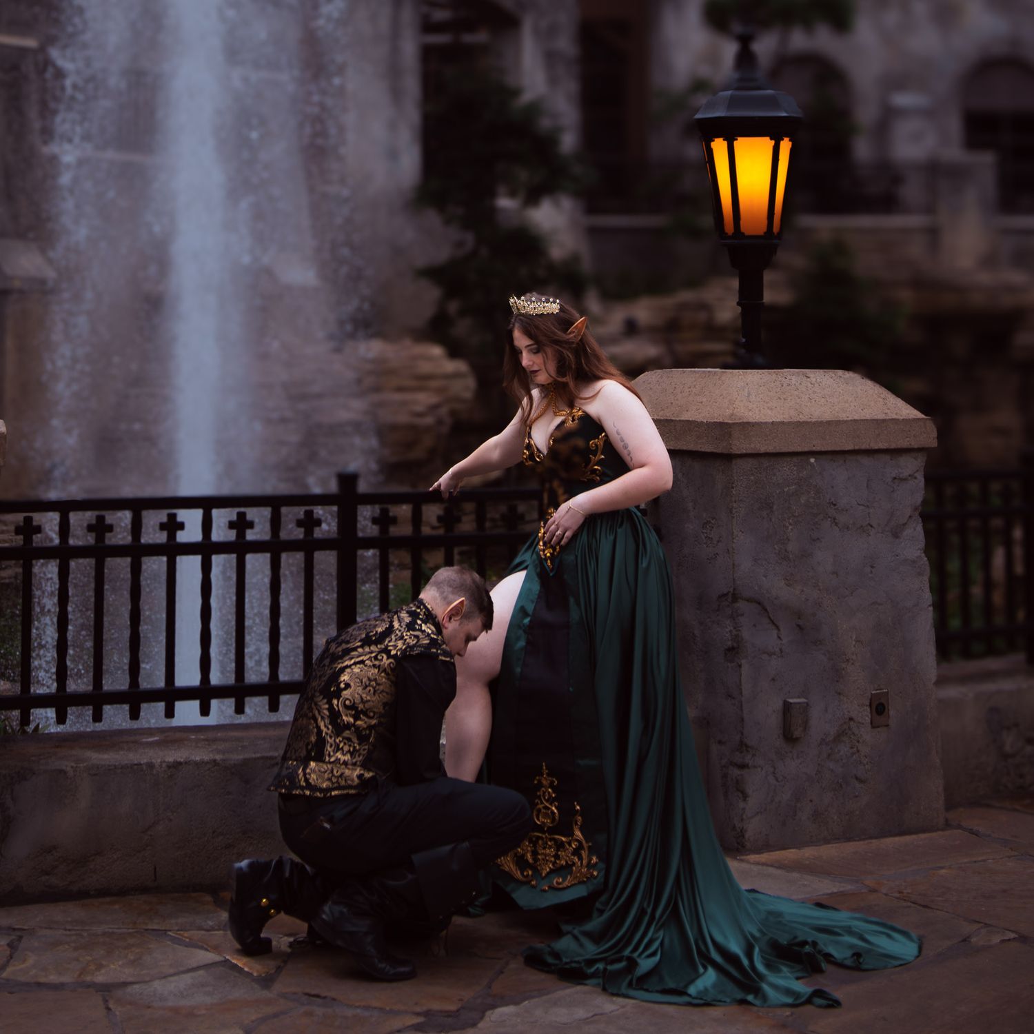 A man in a gold vest knees and helps a woman in a green elven gown adjust her shoe at a Gaylord Texan event, with a fountain background.