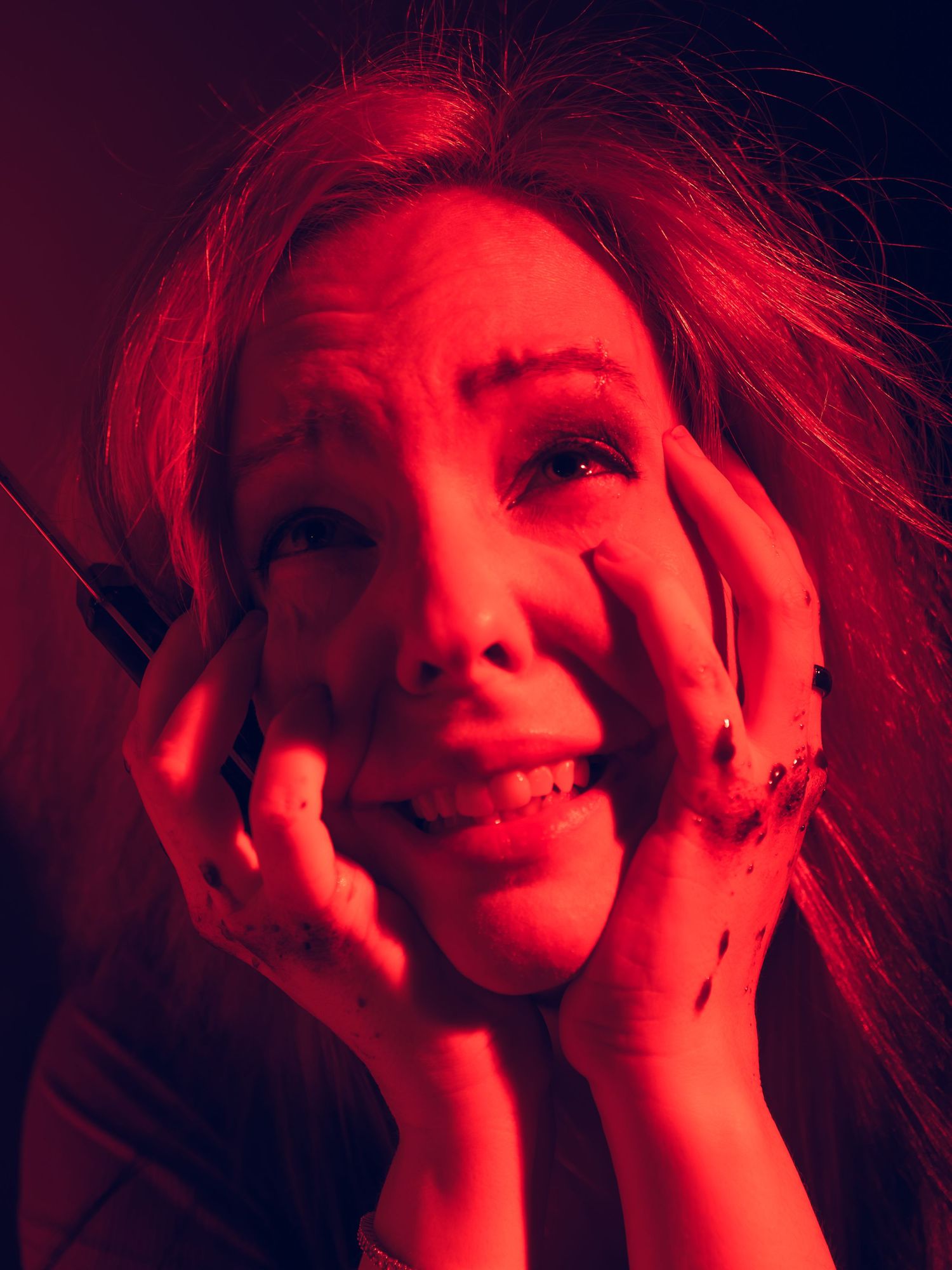 Woman with dramatic red lighting, disheveled hair, and bloody knuckles crying with hands on face.