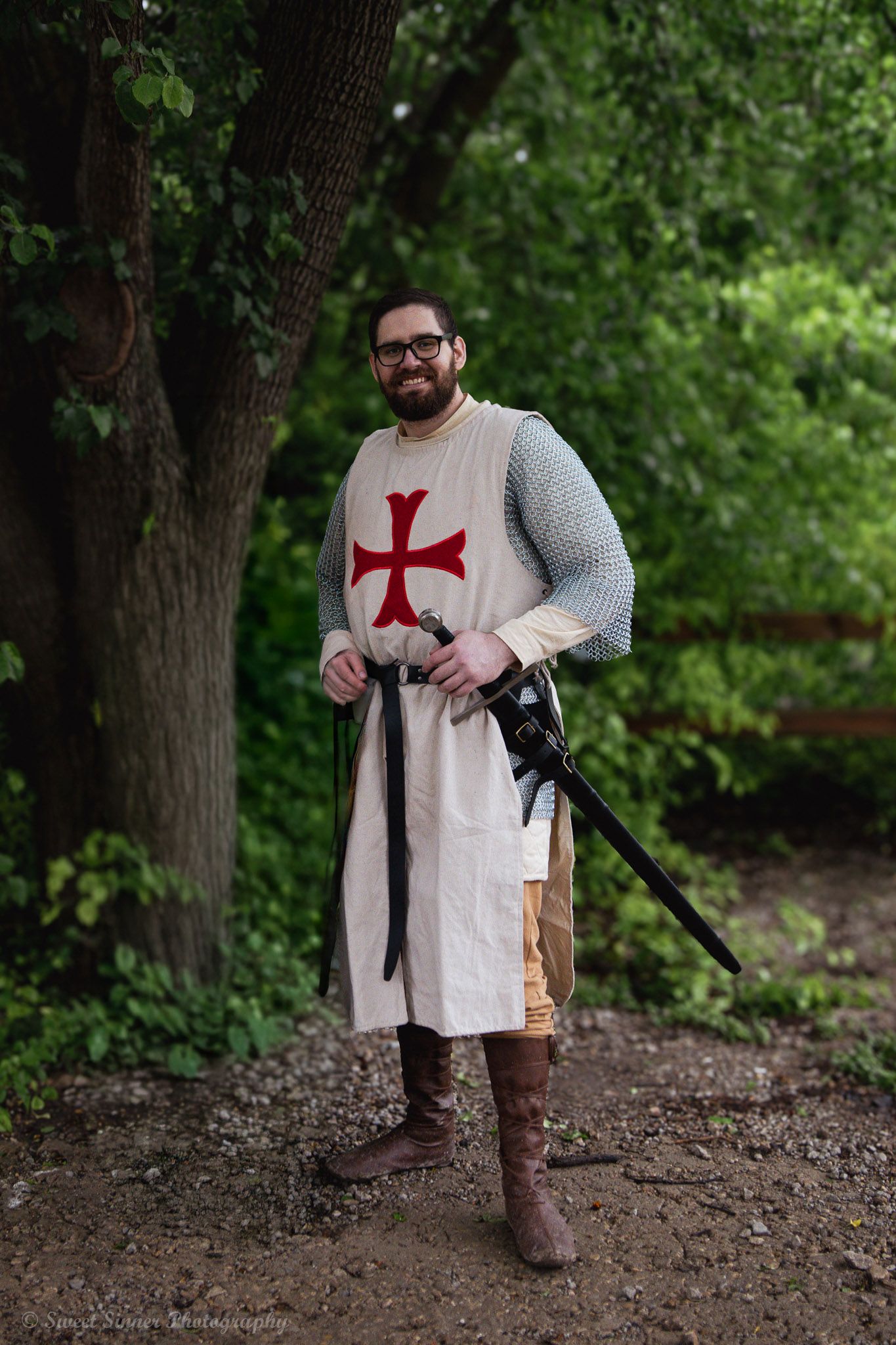 Man in Crusader knight costume with red cross tabard, chainmail, and sword posing outdoors.