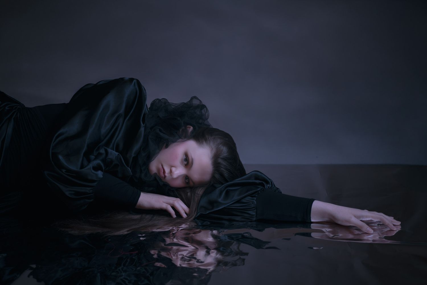 A woman in black clothing lies dramatically on a reflective dark surface in a moody, artistic portrait.