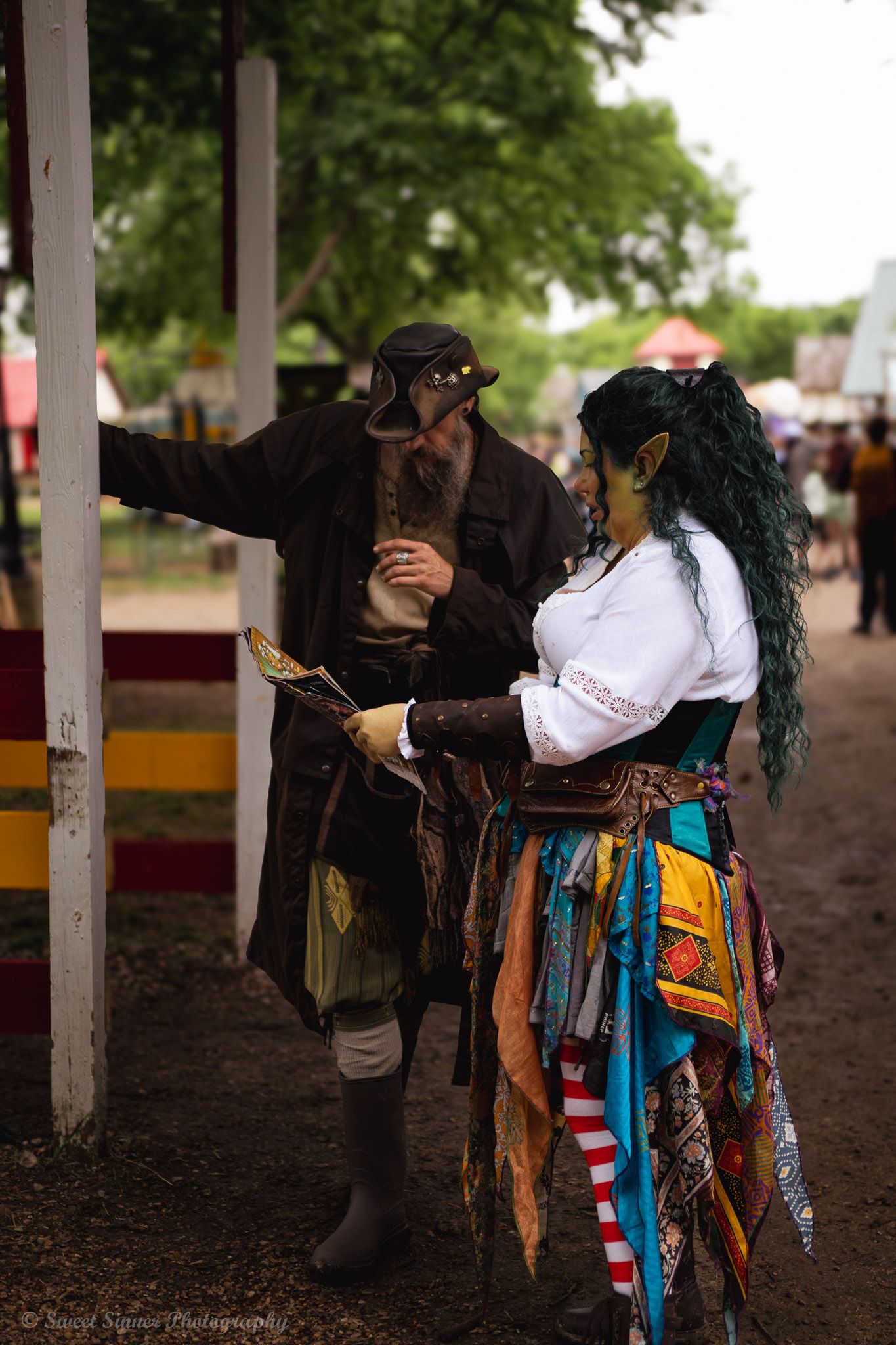 Two costumed Renaissance faire performers, a pirate and a goblin, interact at an outdoor festival.