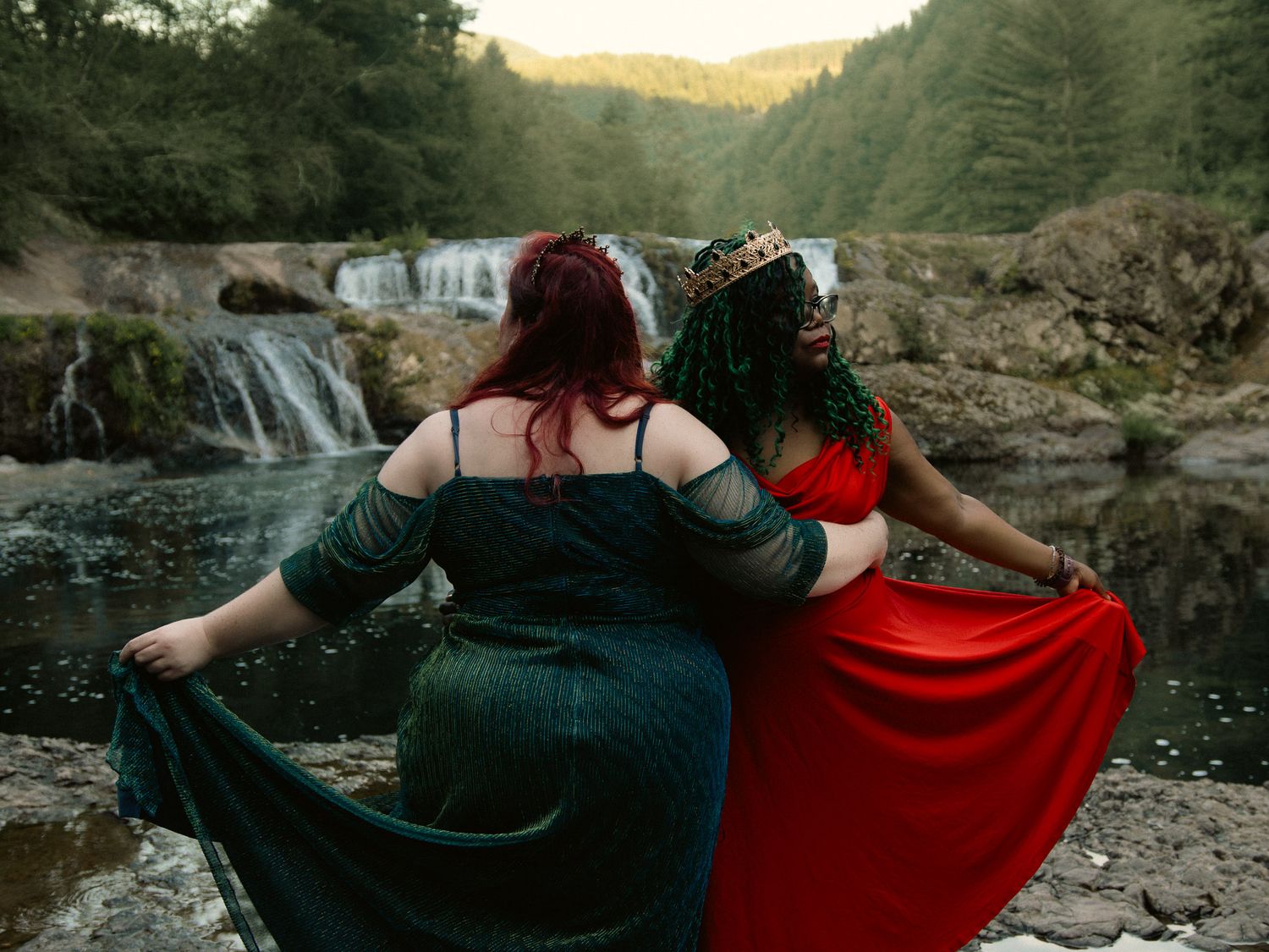 Two women in elegant red and green gowns pose regally before a scenic waterfall in a lush forest.