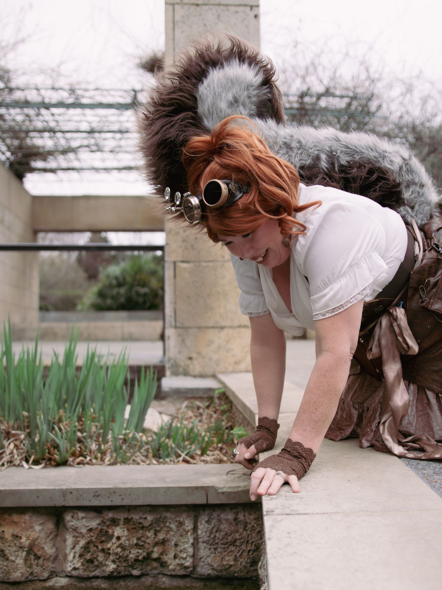 Woman in steampunk costume with goggles and fur accessories leaning over a garden pond outdoors.