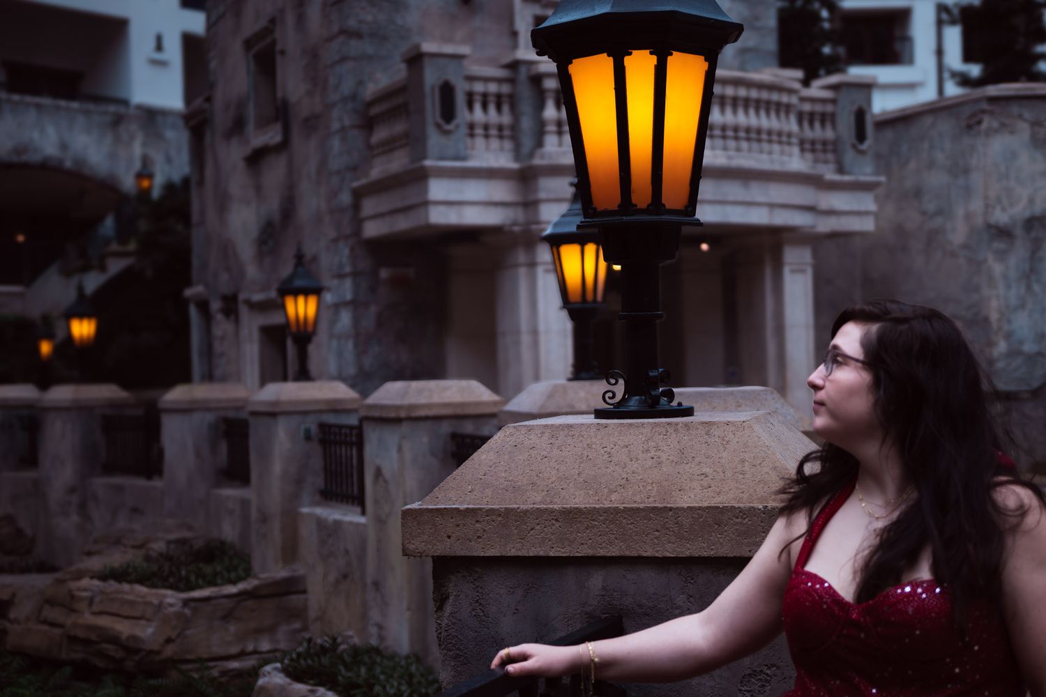 Woman in red dress gazes up at glowing vintage lantern at dusk near stone architecture.
