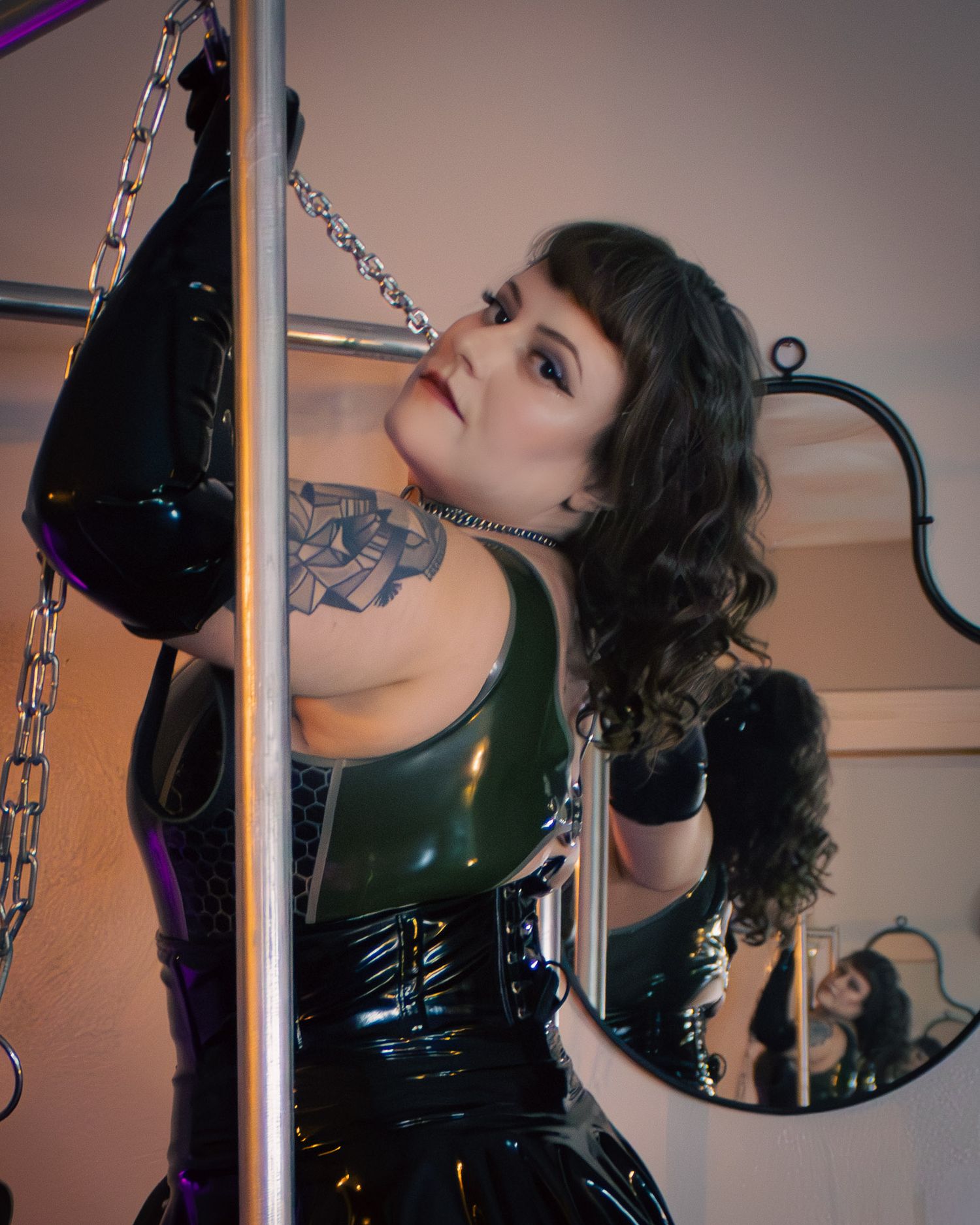 A woman in a dark green and black latex corset outfit with a shoulder tattoo poses near silver poles and chains in a boudoir setting.