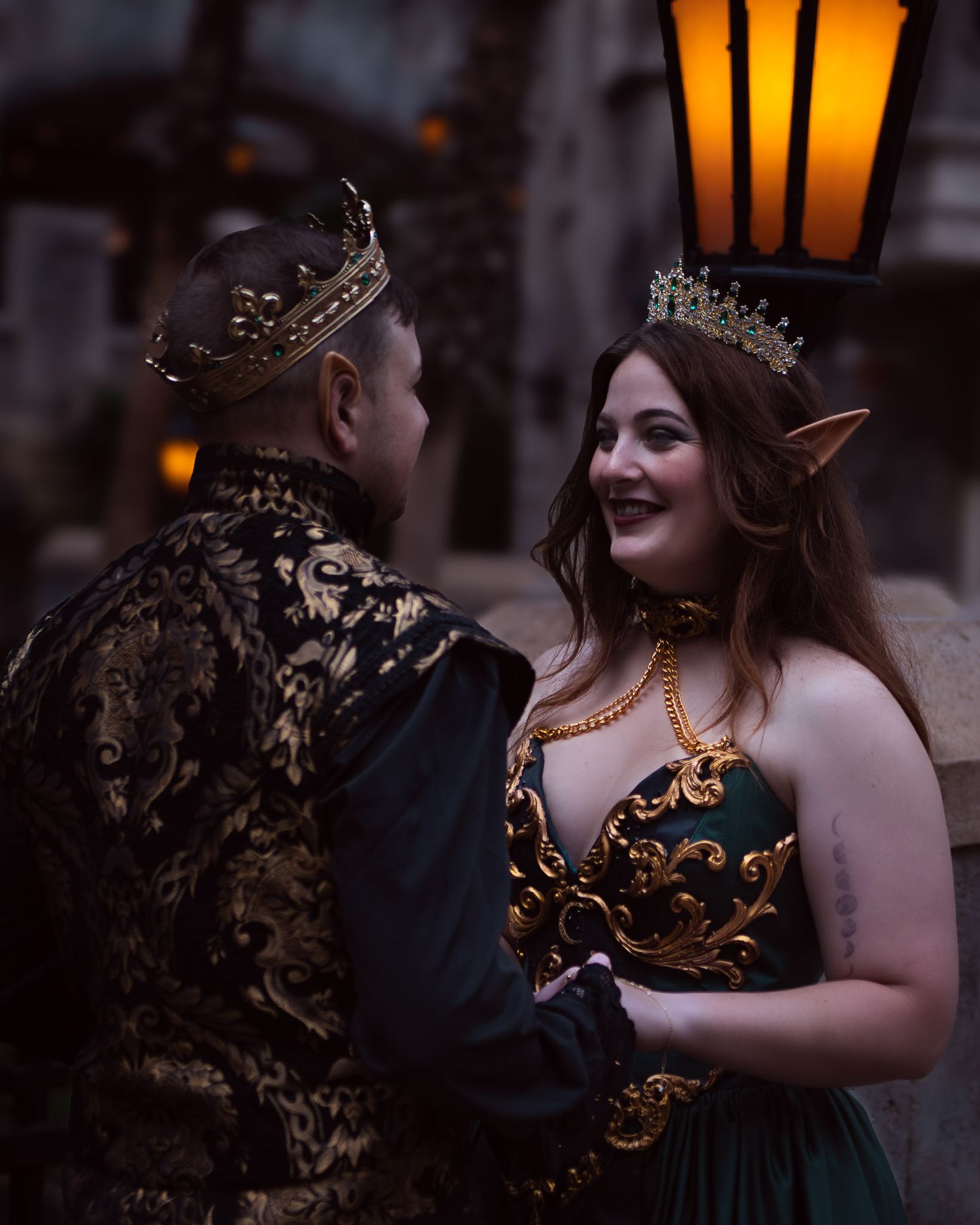 A couple in fantasy medieval costumes, man in crown and brocade jacket, woman with elf ears and tiara, pose under lantern light.