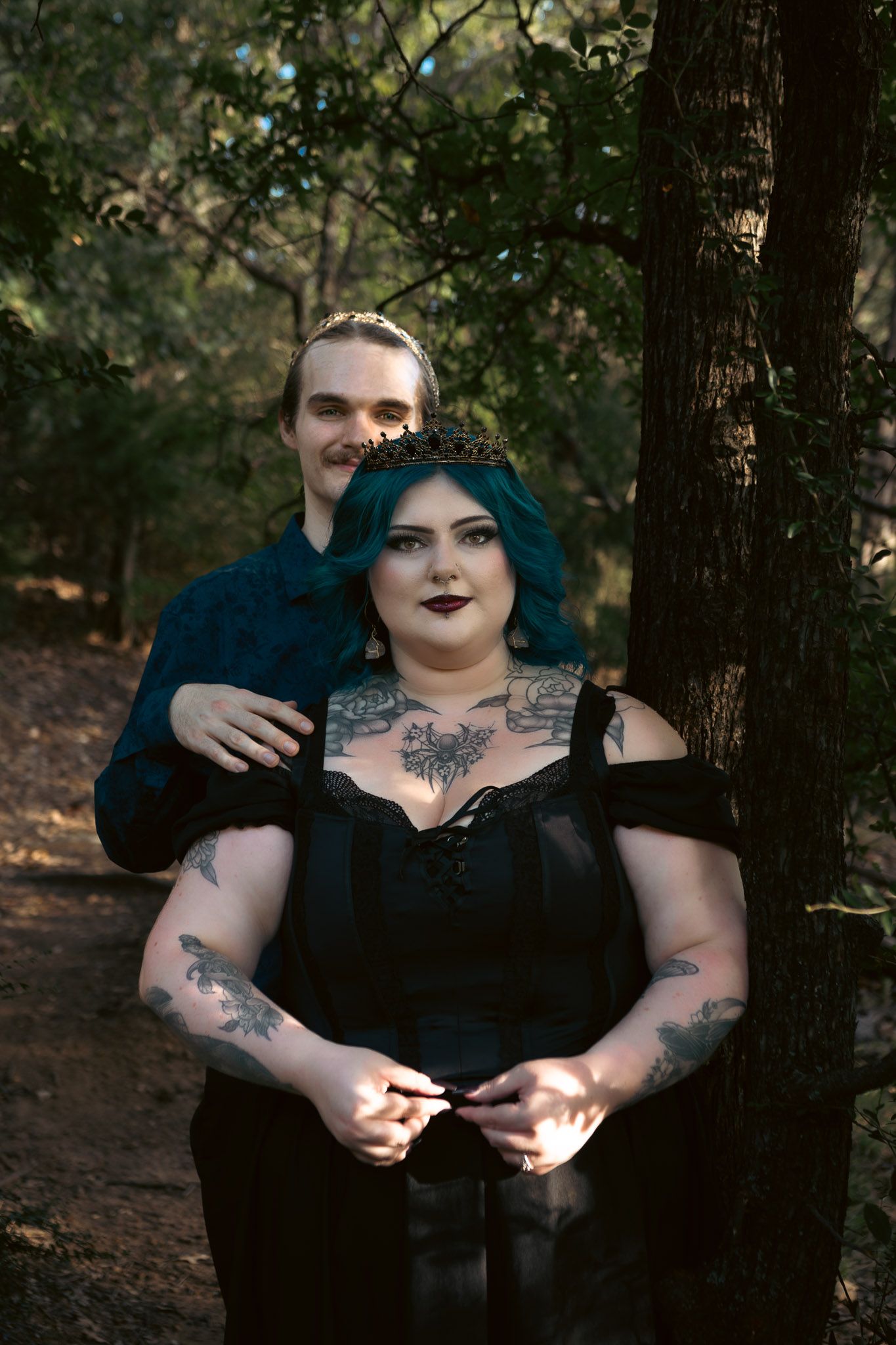 Goth couple in forest, woman with teal hair, tattoos, black dress, crown, man in blue shirt embracing her by tree.