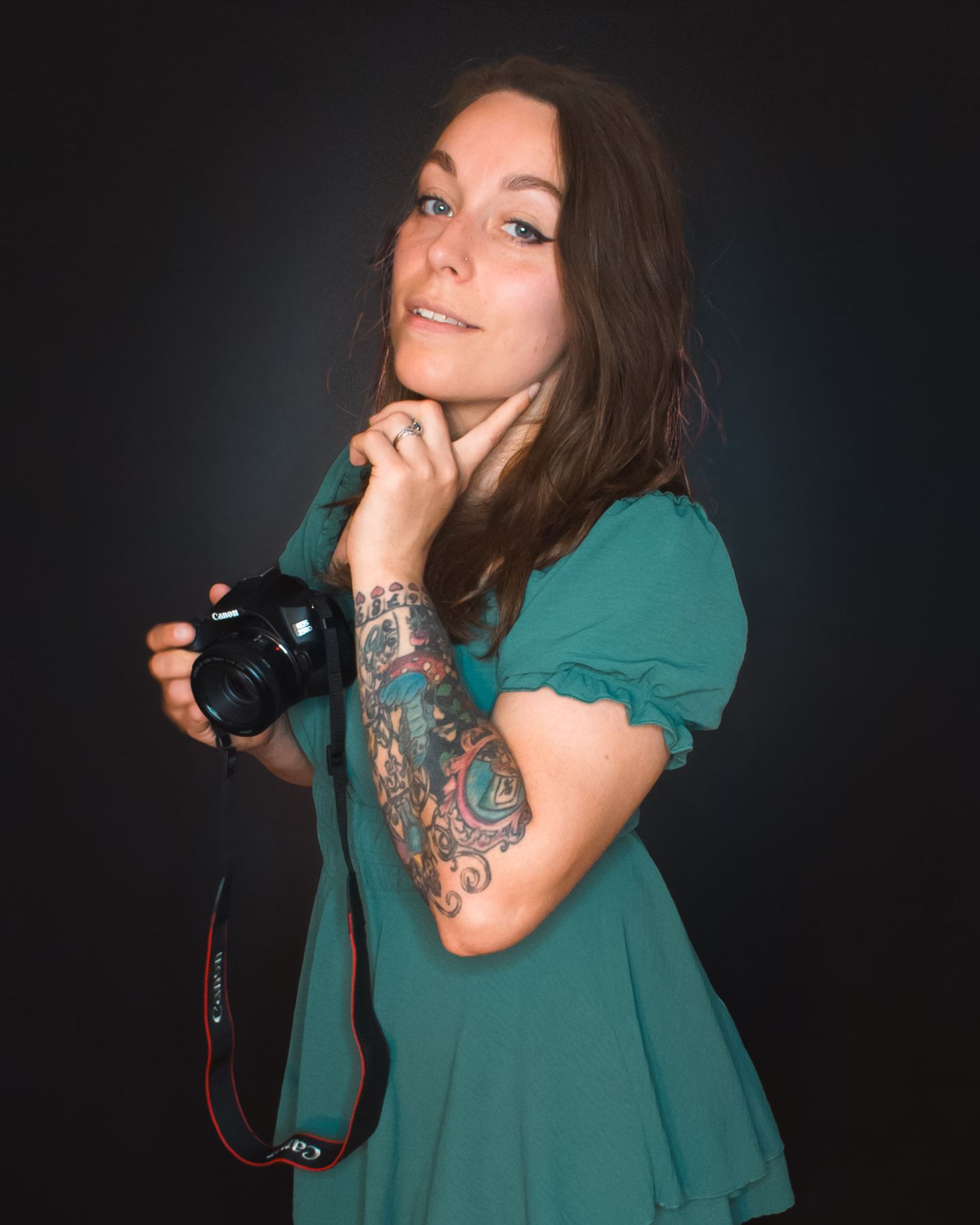 Woman in teal dress holding Canon camera with colorful tattoo sleeve, posing against dark background.
