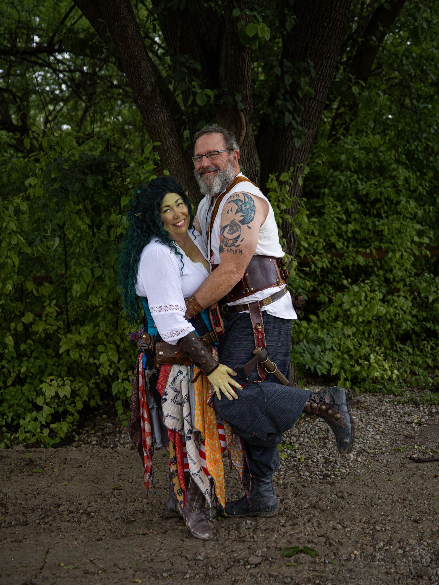 Couple in fantasy cosplay costumes pose outdoors, goblin woman with green hair and green makeup, man with tattoos and leather gear.