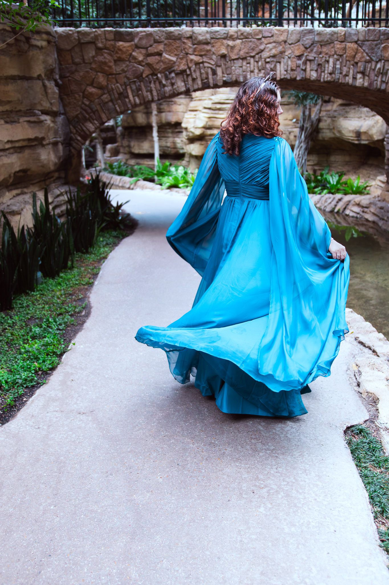 Woman in flowing teal gown walks along stone path toward rustic bridge in lush garden setting.