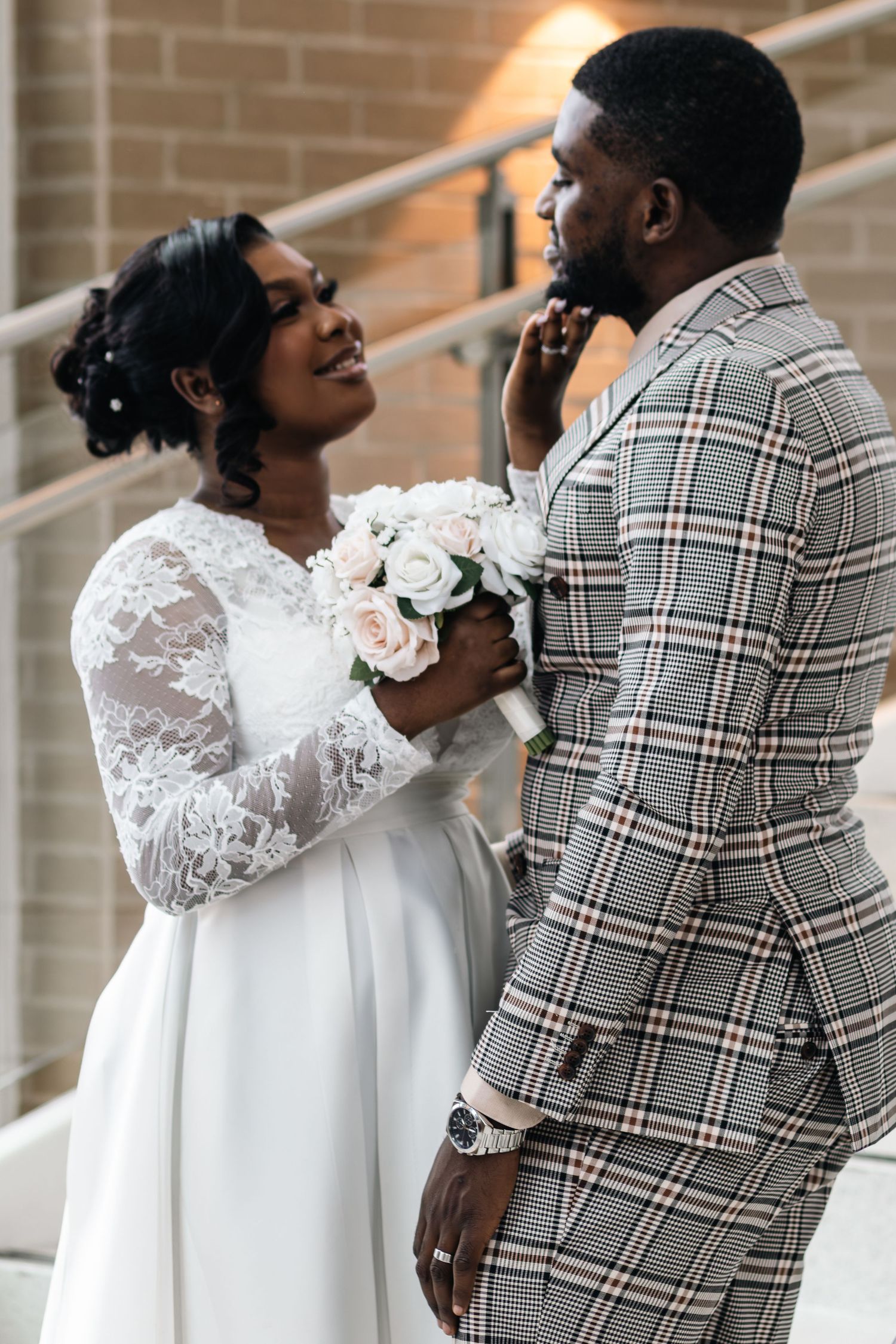 A bride in white lace gown holds a rose bouquet while touching groom's face in plaid suit on wedding day.