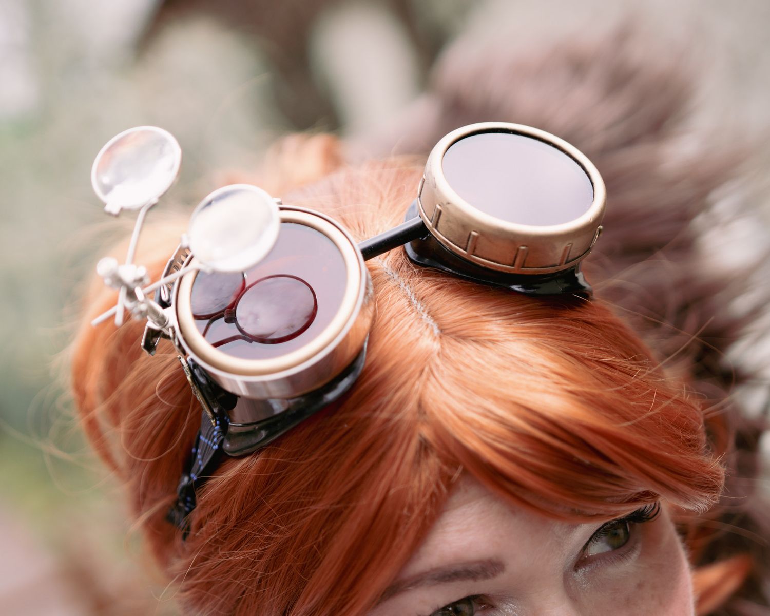 Woman with red hair wearing steampunk goggles with multiple lenses outdoors.