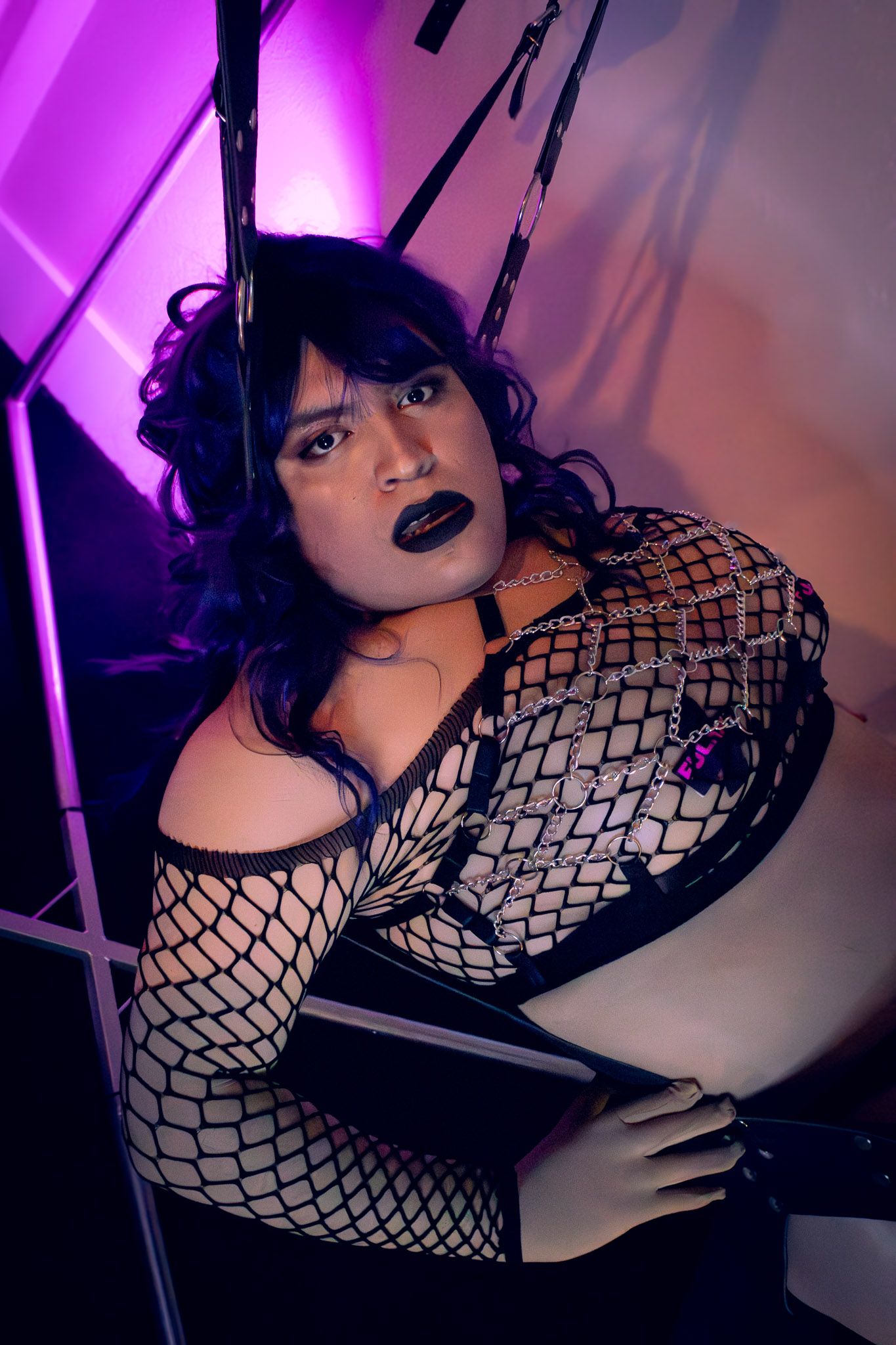 Person in fishnet outfit with dark makeup poses under purple neon lights in edgy gothic style fashion shoot.