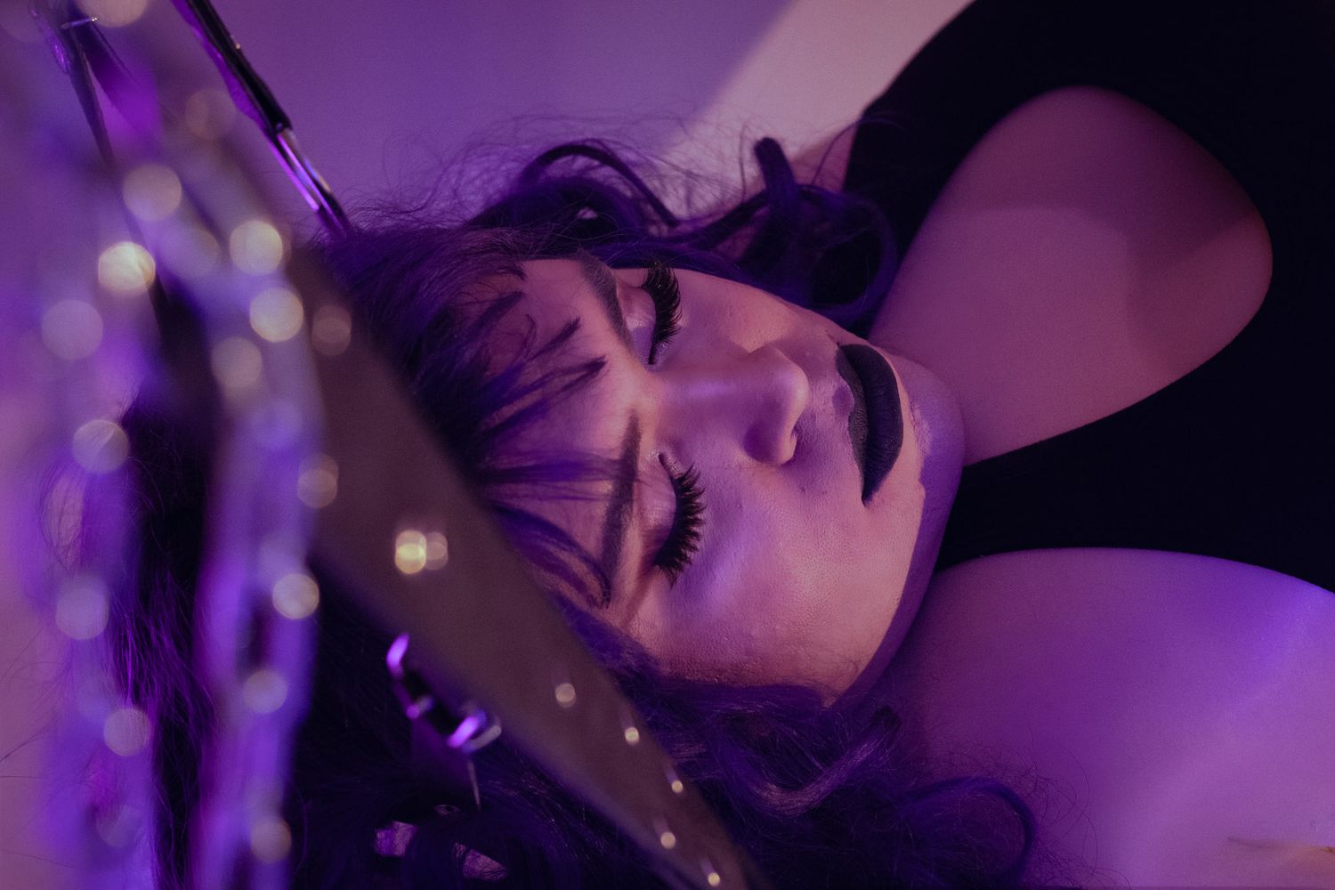 Woman with purple hair and dramatic makeup lying under violet lighting with bokeh effects.