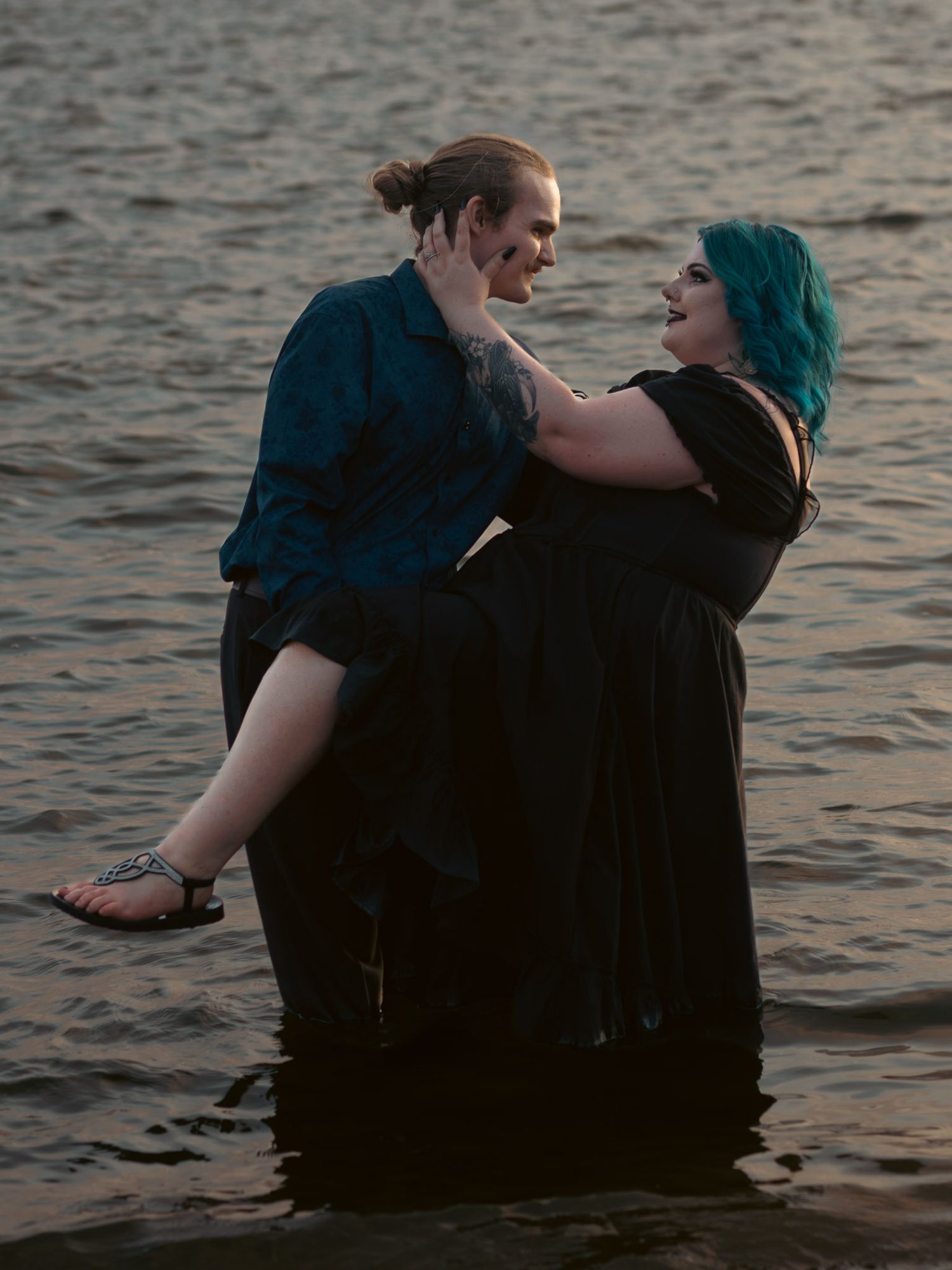Couple embracing romantically while standing in water, woman with teal hair in black dress being held intimatelty by partner in blue shirt.