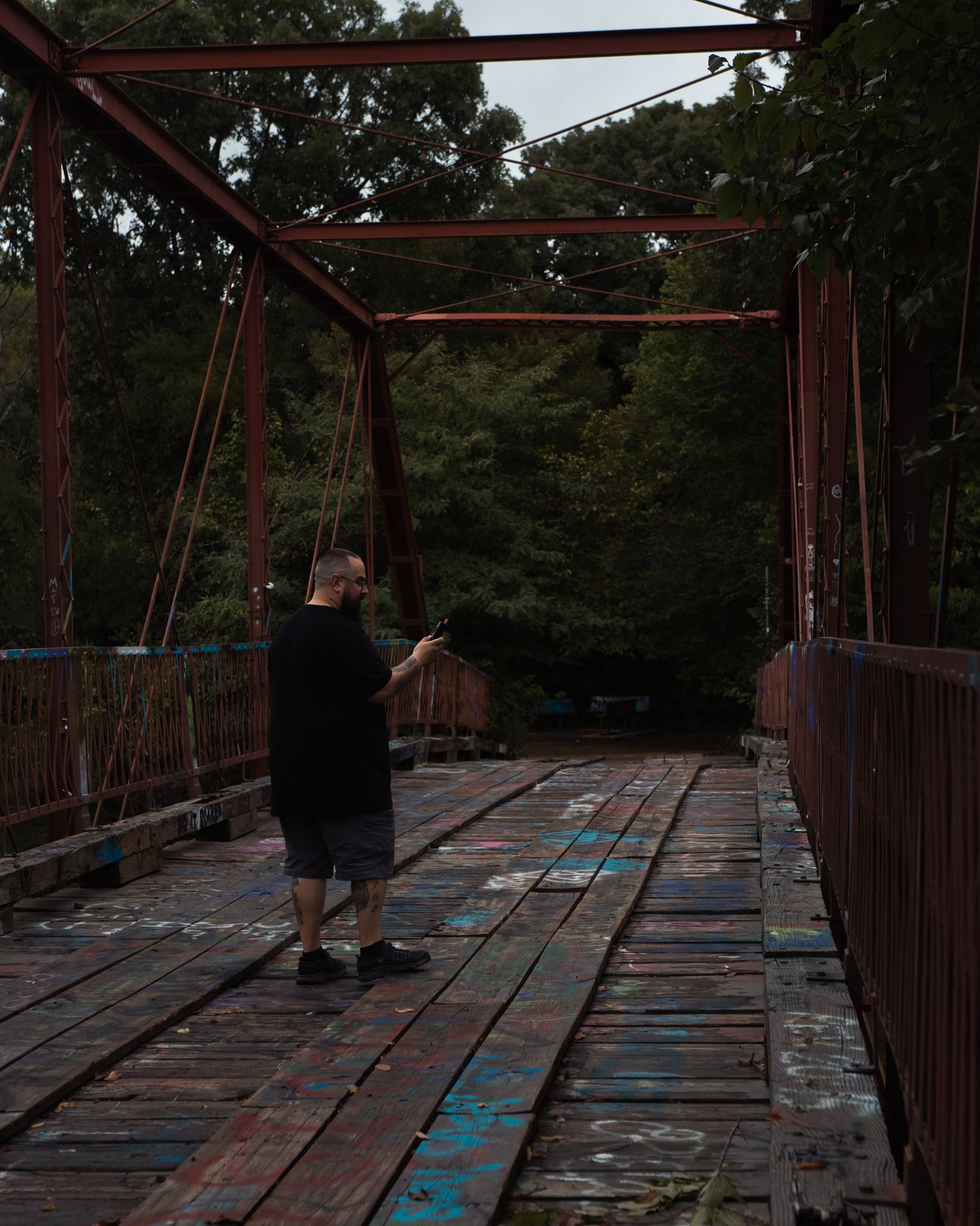 Person standing on a weathered, graffiti-covered iron bridge surrounded by lush green trees, using ghost hunting equipment.