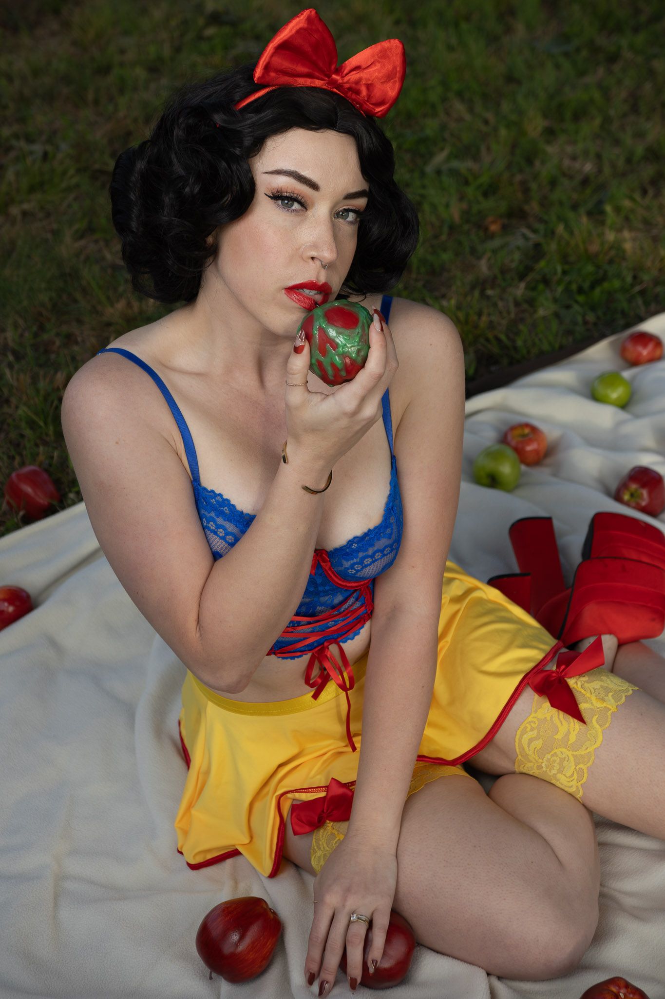Woman in a Snow White-inspired cosplay with red bow, blue lace top and yellow skirt holds a green apple on a blanket among scattered apples.