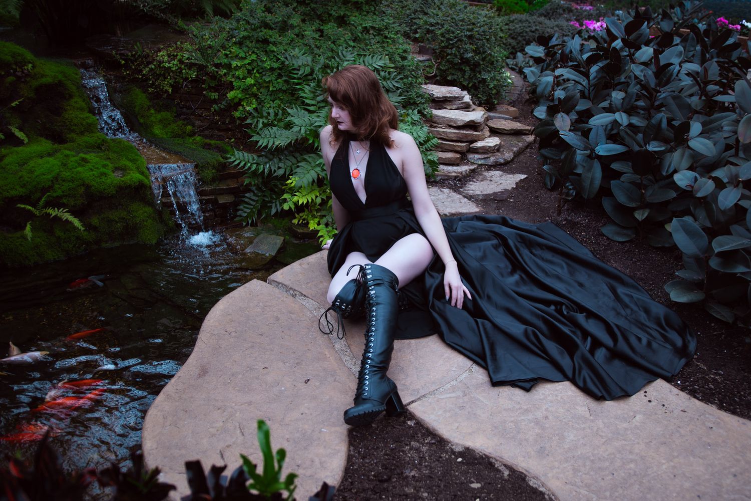 Woman in black gown and boots sitting by a garden pond surrounded by lush greenery and ferns.