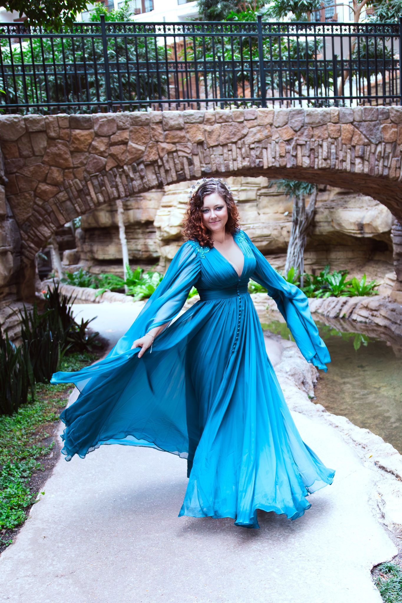 Woman in flowing teal gown twirling near stone bridge in lush garden setting.