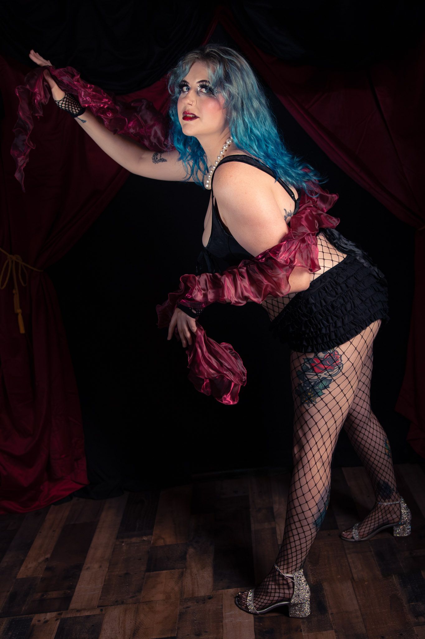 Blue-haired burlesque performer in black costume with red ruffles poses dramatically on a dark stage.