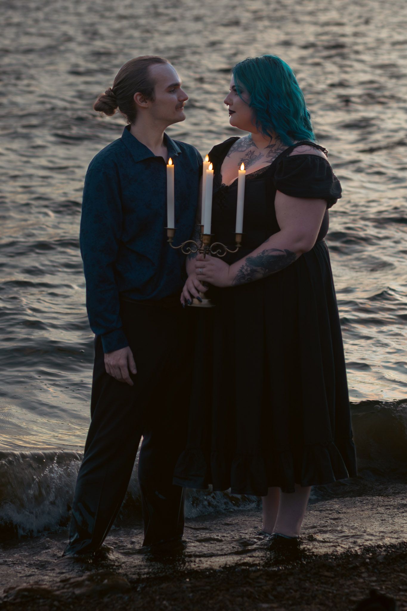 Couple holding candelabra by the lake at dusk, woman with teal hair in black dress, man in blue shirt.