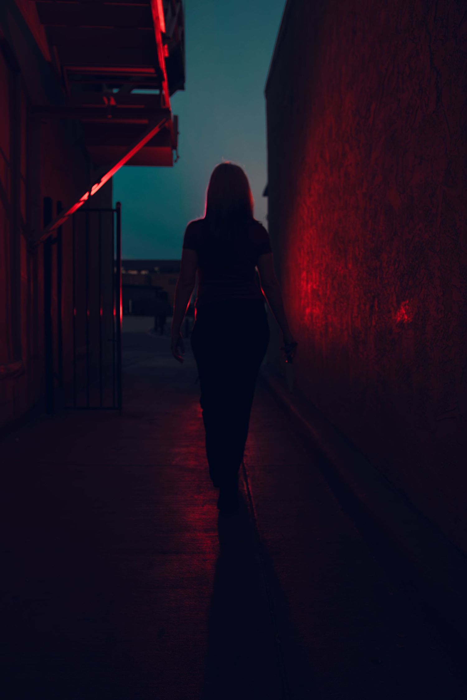 Silhouette of a woman walking alone through a dark alley illuminated by dramatic red neon lights at night.
