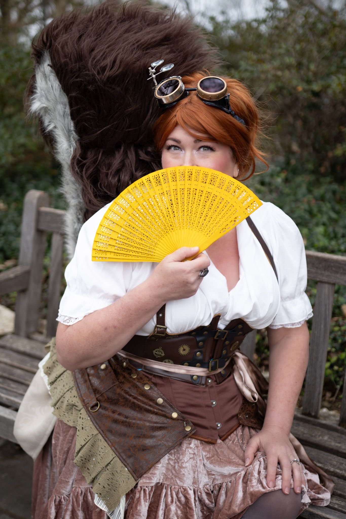 Woman in steampunk costume with corset, goggles, and yellow fan at outdoor event.