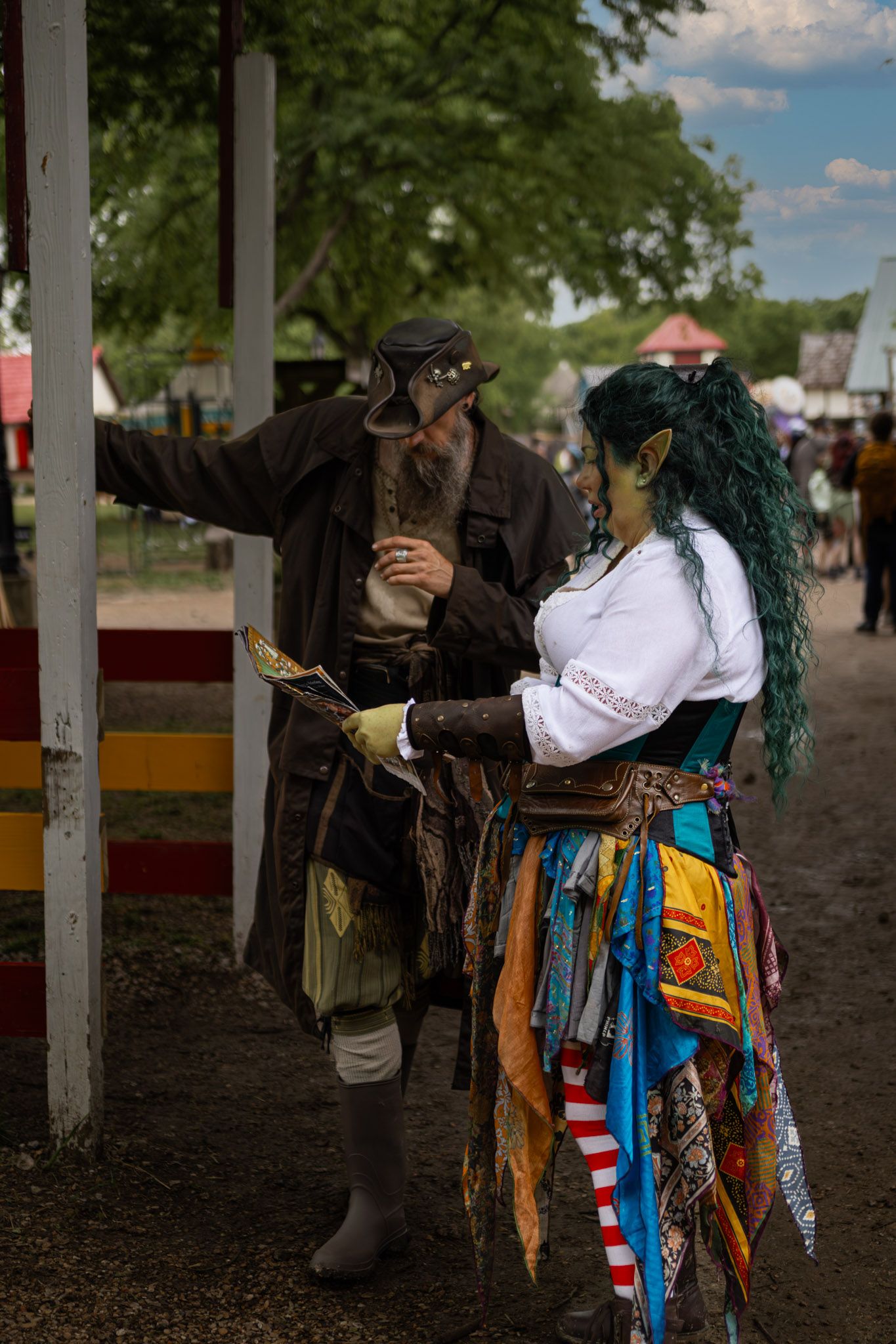 Two cosplayers in fantasy costumes at a Renaissance faire, one dressed as a pirate, the other as a green-haired goblin.