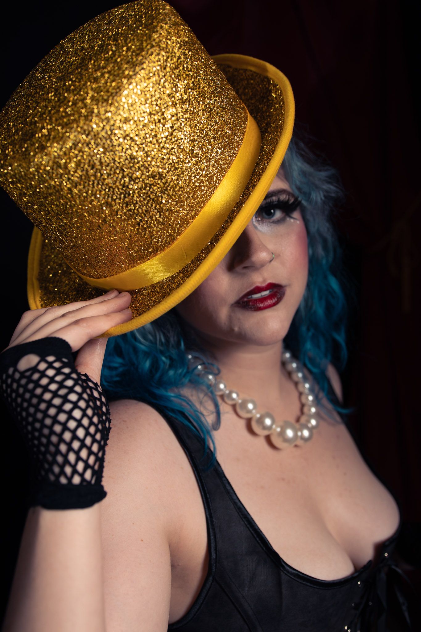Woman in gold glitter top hat, blue hair, pearl necklace, black corset and fishnet gloves posing dramatically.