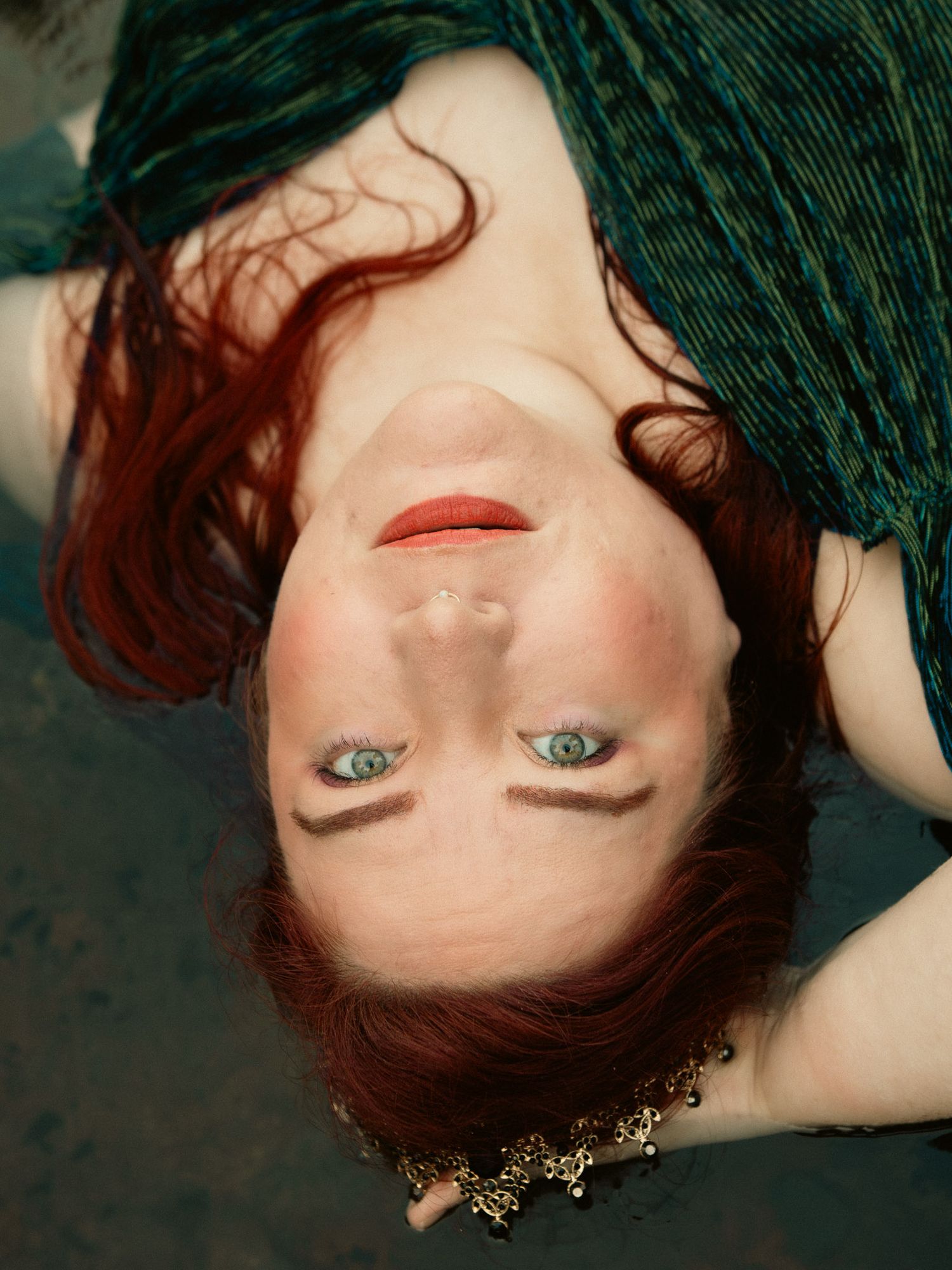 A woman with red hair and green eyes lying upside down in a clear riverbed, wearing a green top and gold necklace.