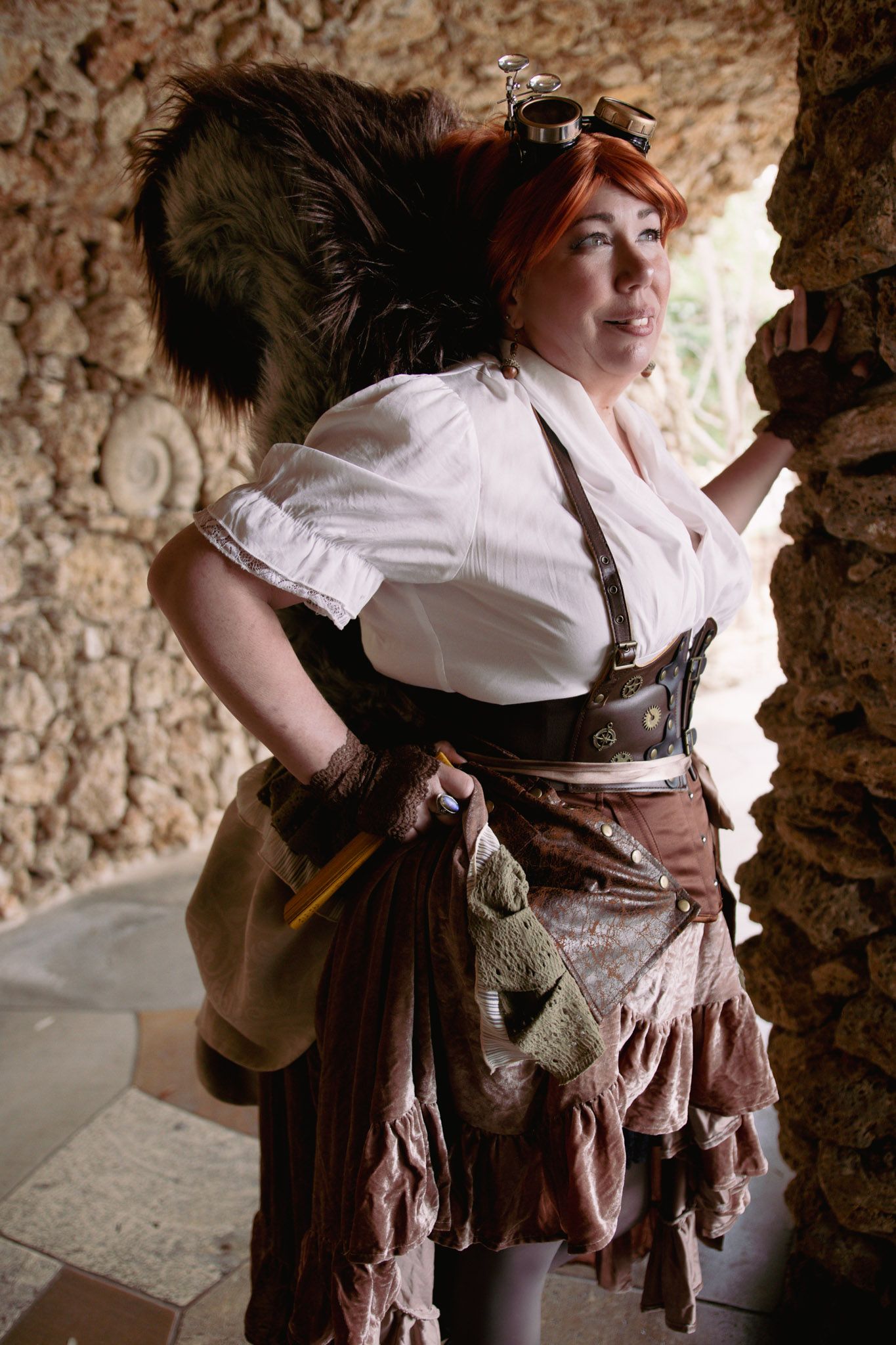 Woman in steampunk Squirrel Girl costume with red hair, goggles, corset, and layered skirt poses by stone wall.