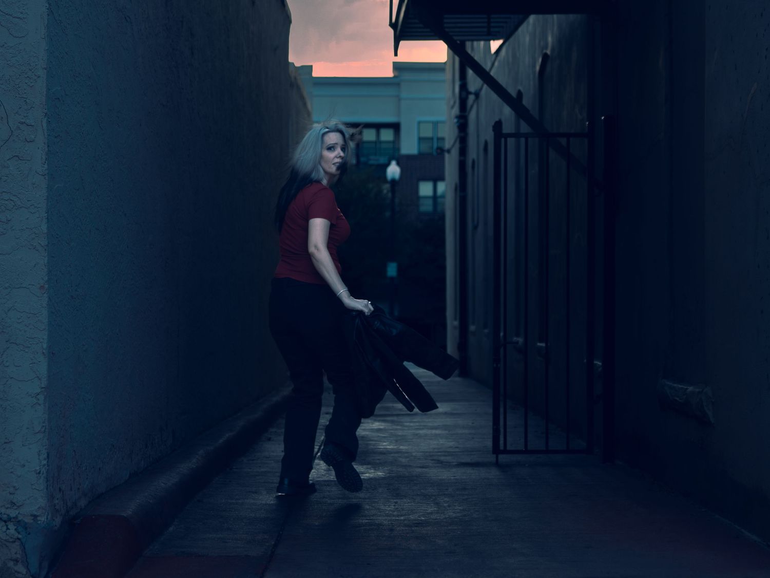 Woman with white hair in red shirt runs in dark alley at dusk, holding black leather jacket near iron gate.