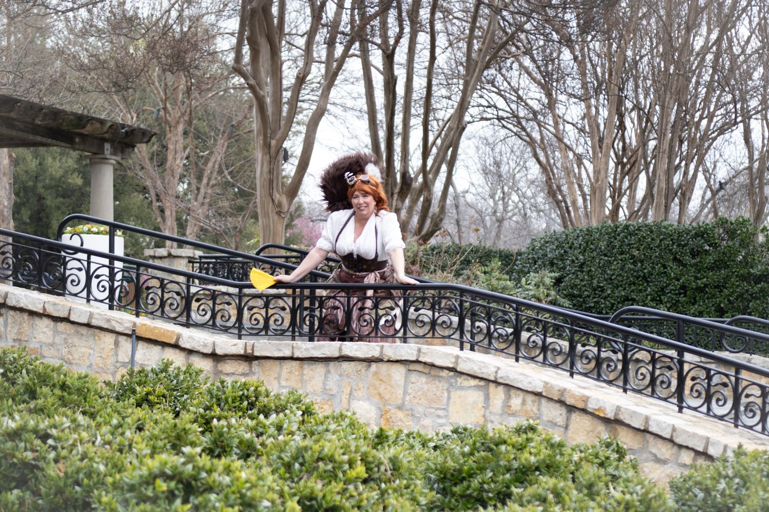 Woman in vintage outfit standing on stone terrace with iron railings surrounded by lush garden trees.