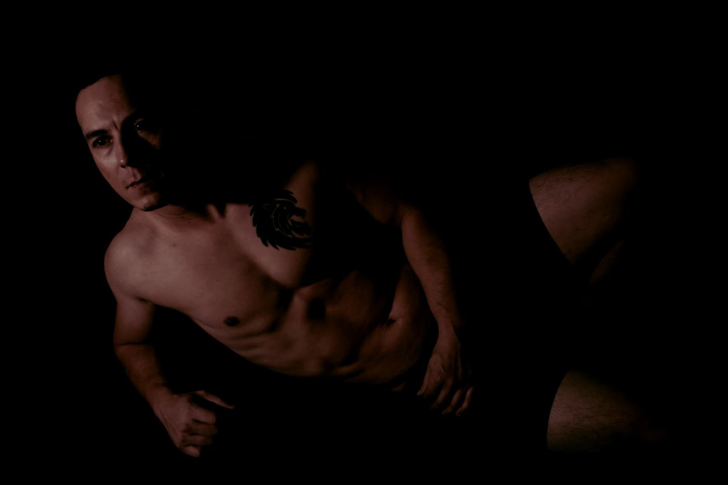 Dramatic low-key portrait of a muscular shirtless man posing in dark moody lighting.