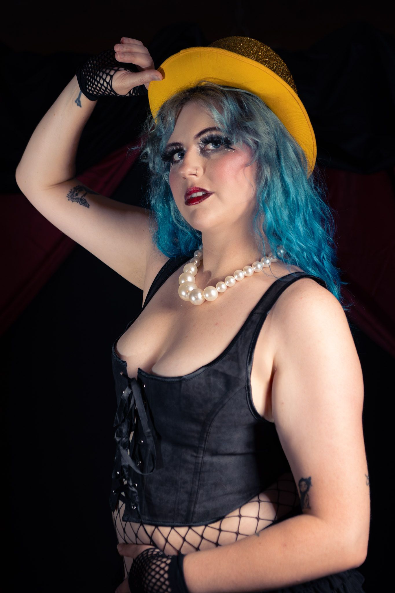 Woman with blue hair wearing a yellow hat, black corset, pearl necklace, and fishnet gloves poses dramatically.