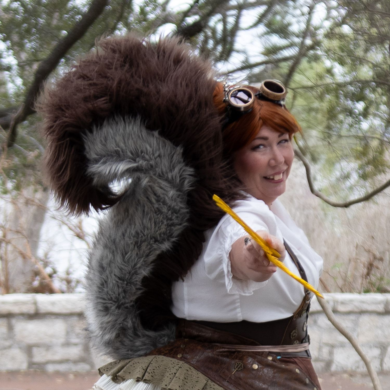 Woman in steampunk squirrel costume with fluffy tail, goggles, and white blouse, posing outdoors.