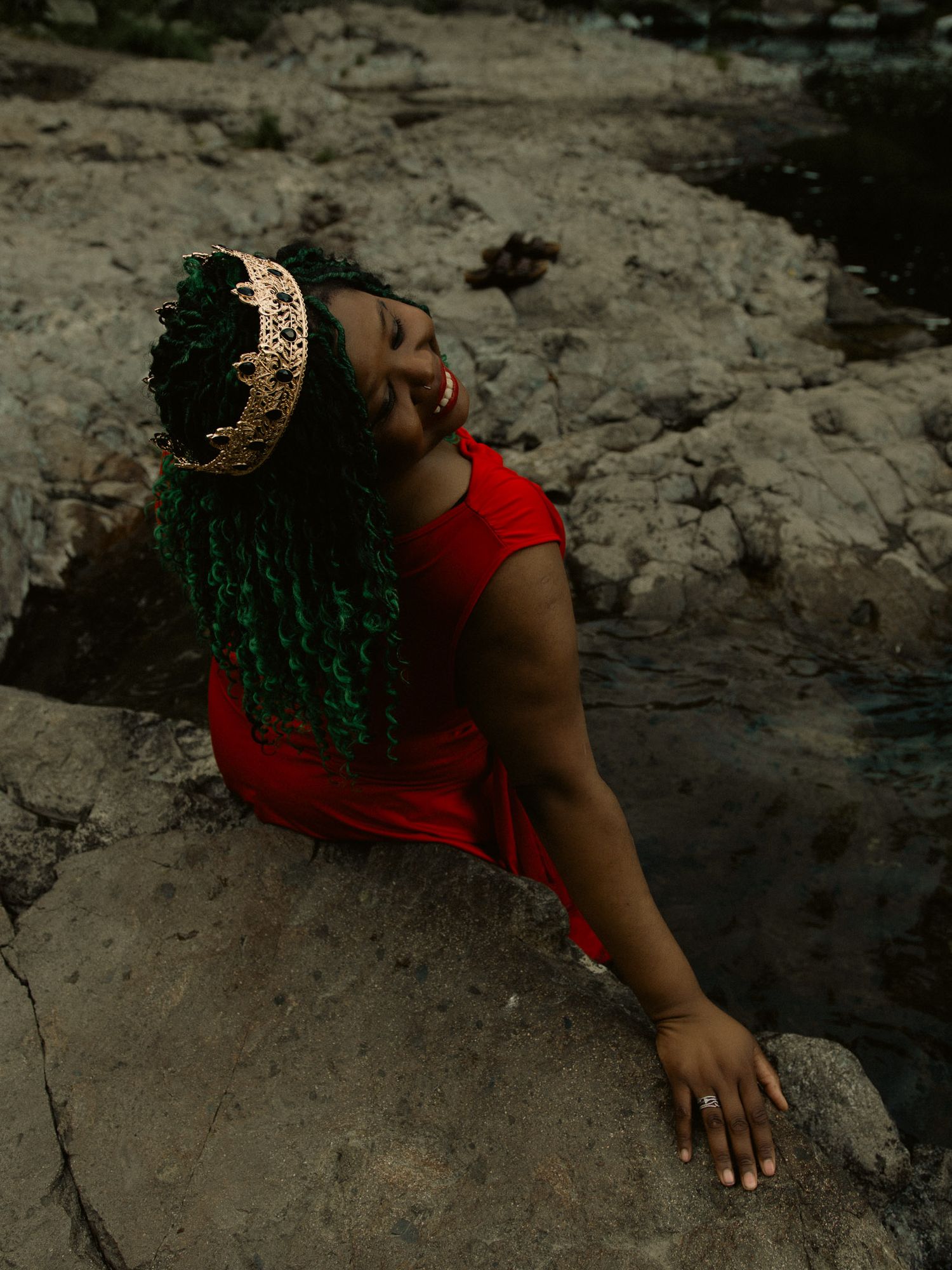 Woman in red dress and gold crown with green hair smiles while sitting on rocky terrain near water.