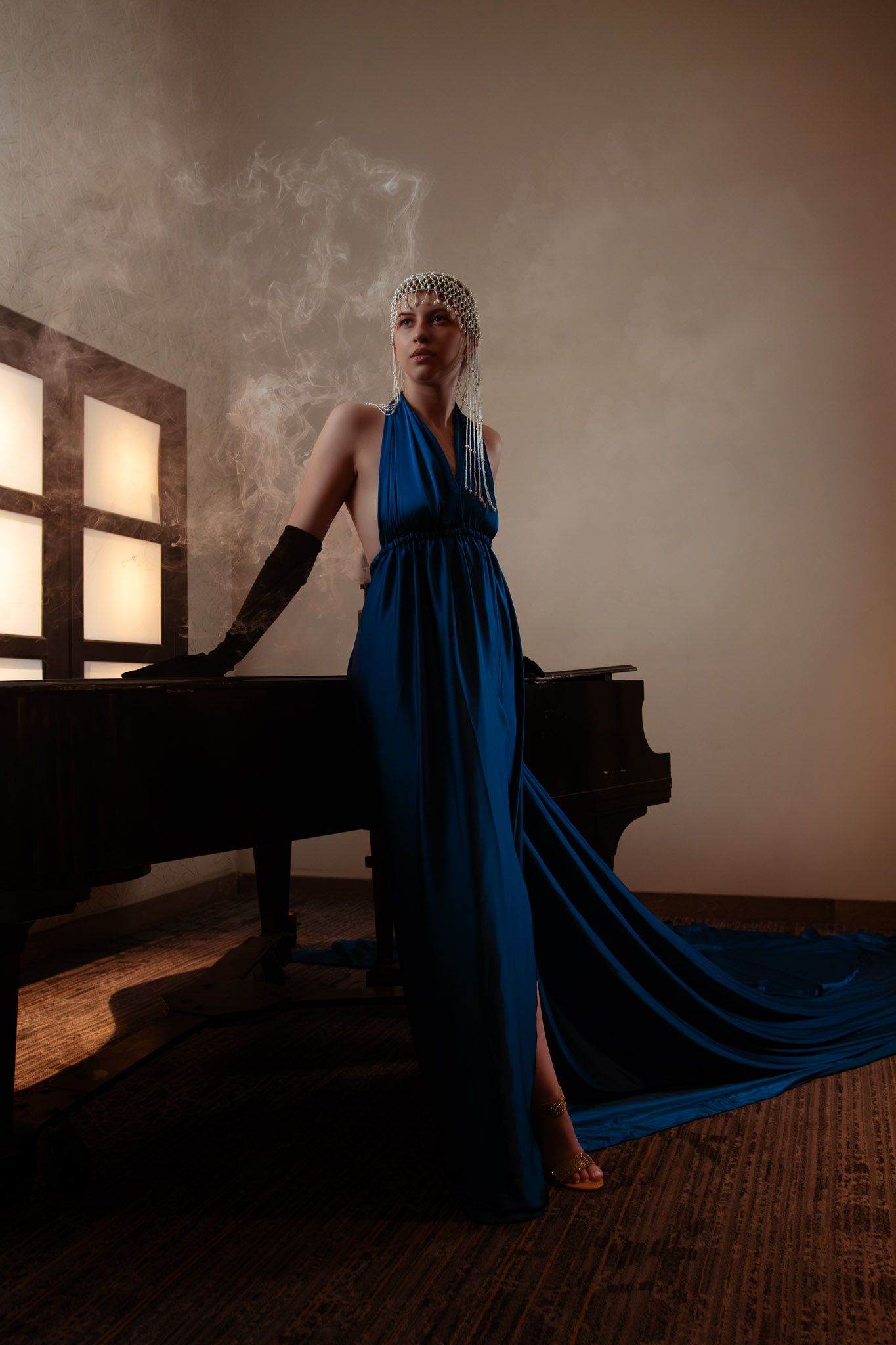 Woman in blue halter gown with black gloves and pearl headpiece poses beside a grand piano in moody lighting.