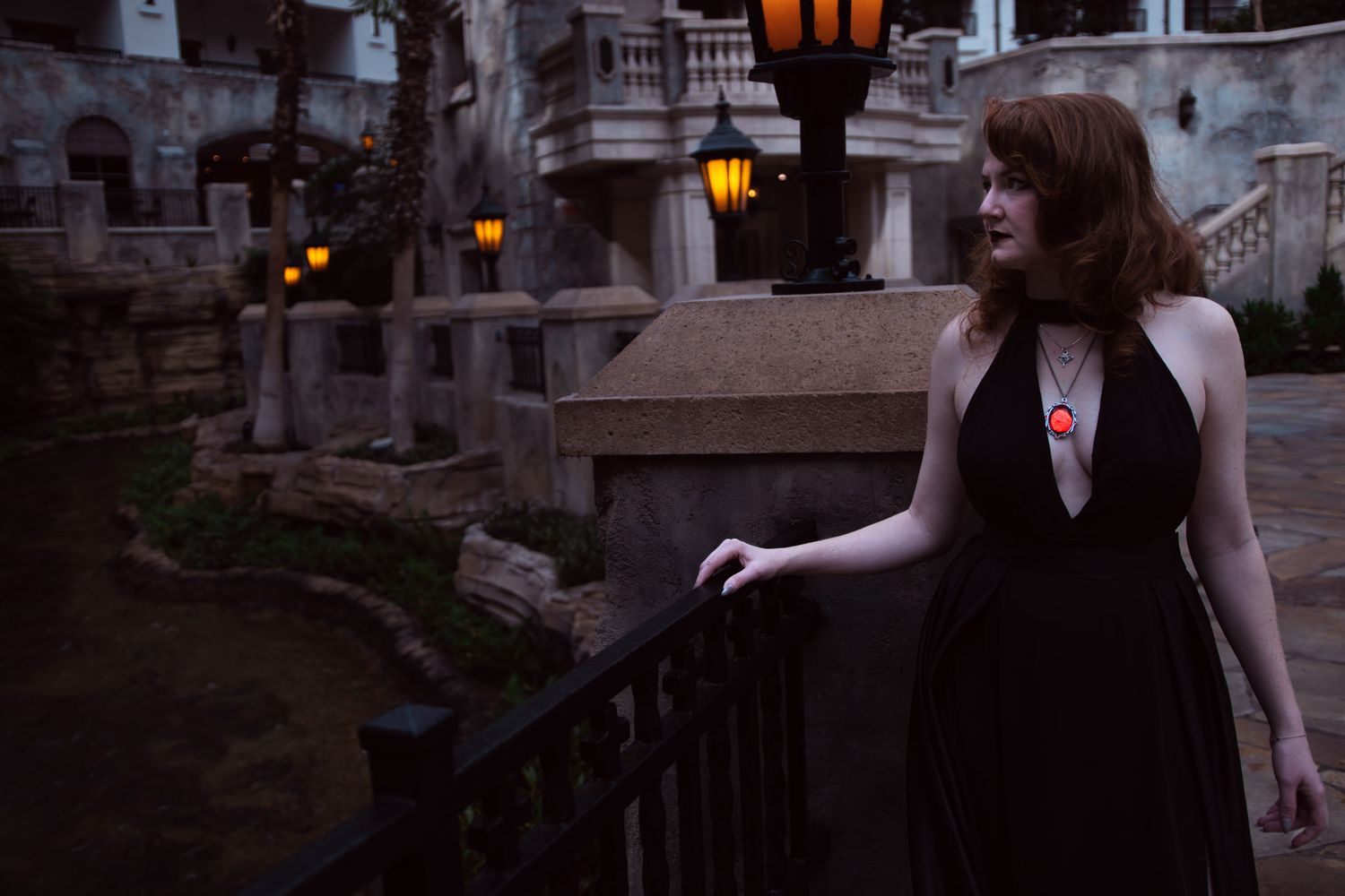 Woman in black halter dress stands by railing at dusk near illuminated stone building.