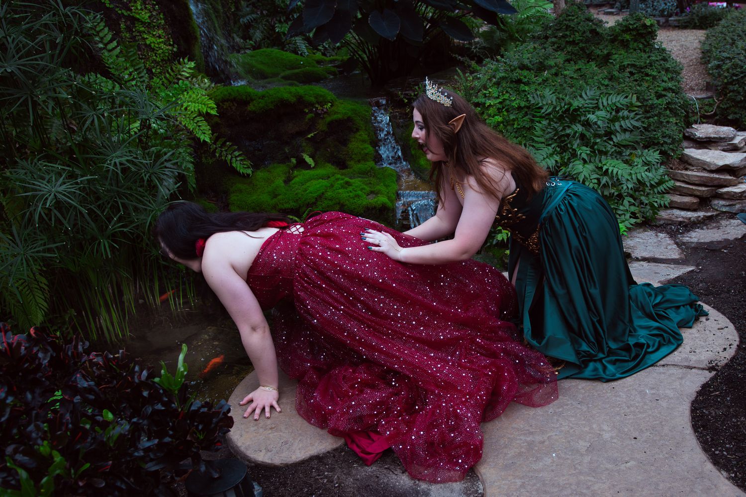 Two women in elegant gowns, one in red and one in green, one pushing the other into the pond in a lush garden setting.