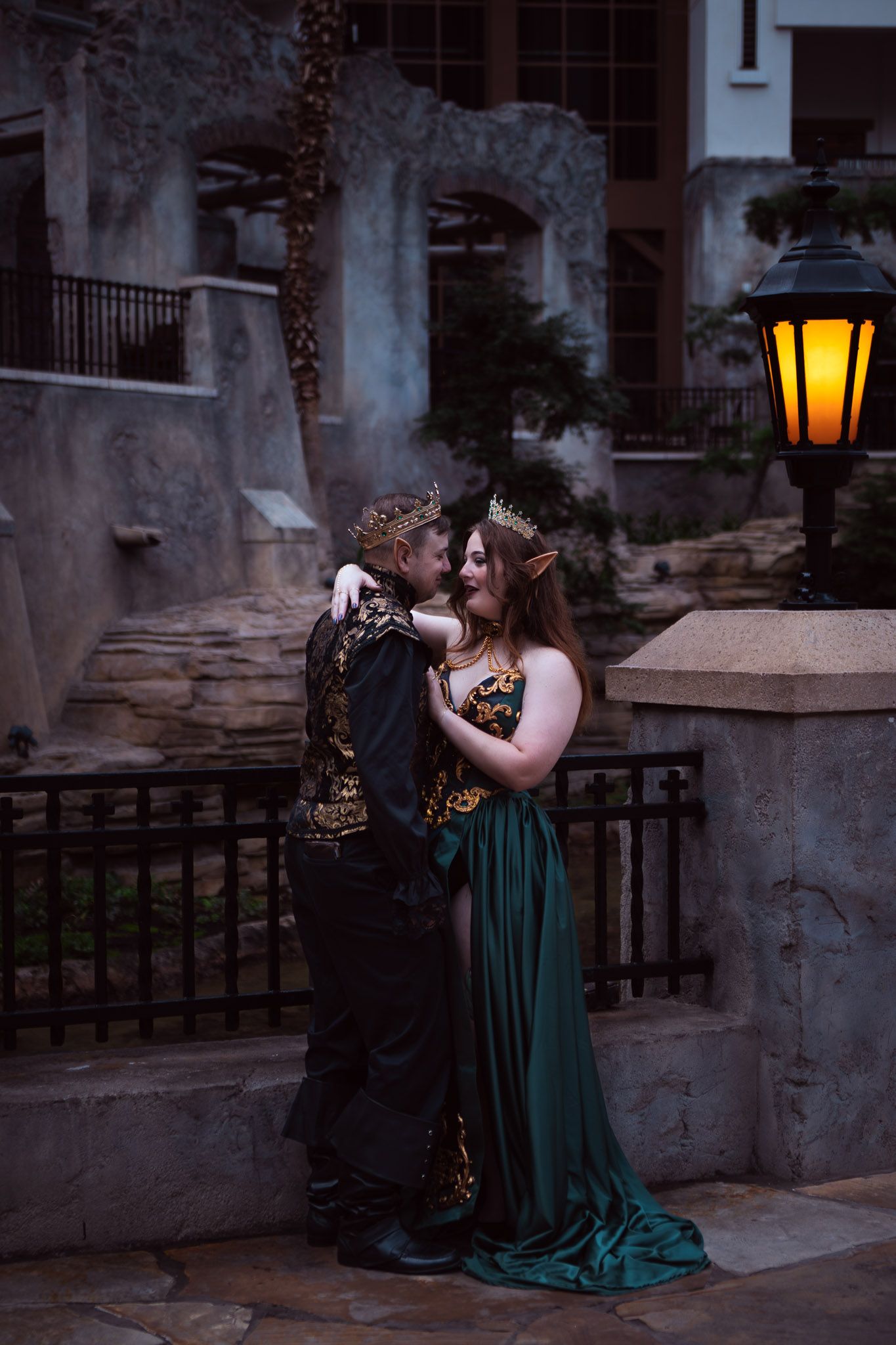 Couple in fantasy elf costumes with crowns embracing near a glowing lantern at dusk.