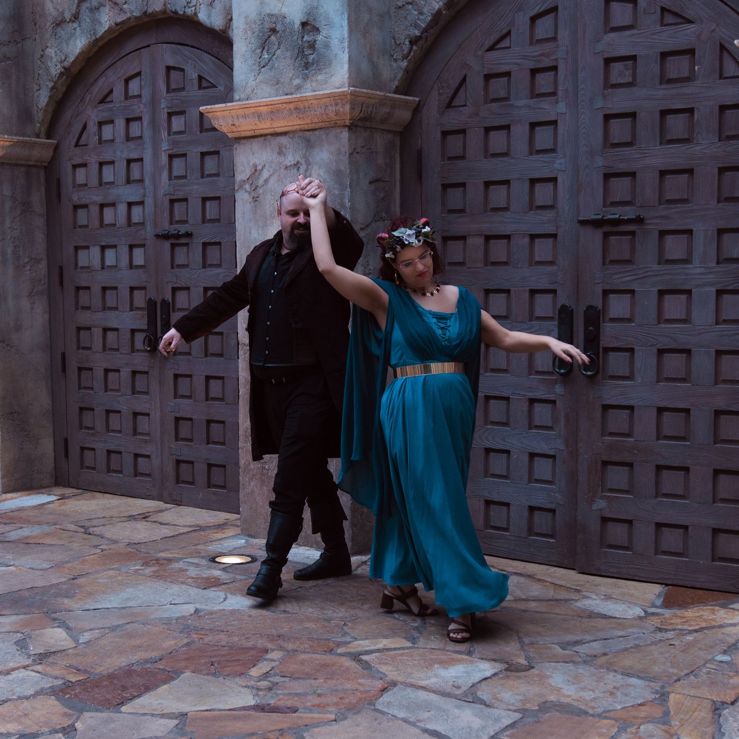 A couple in medieval costumes dancing in front of ornate wooden doors on a stone courtyard.
