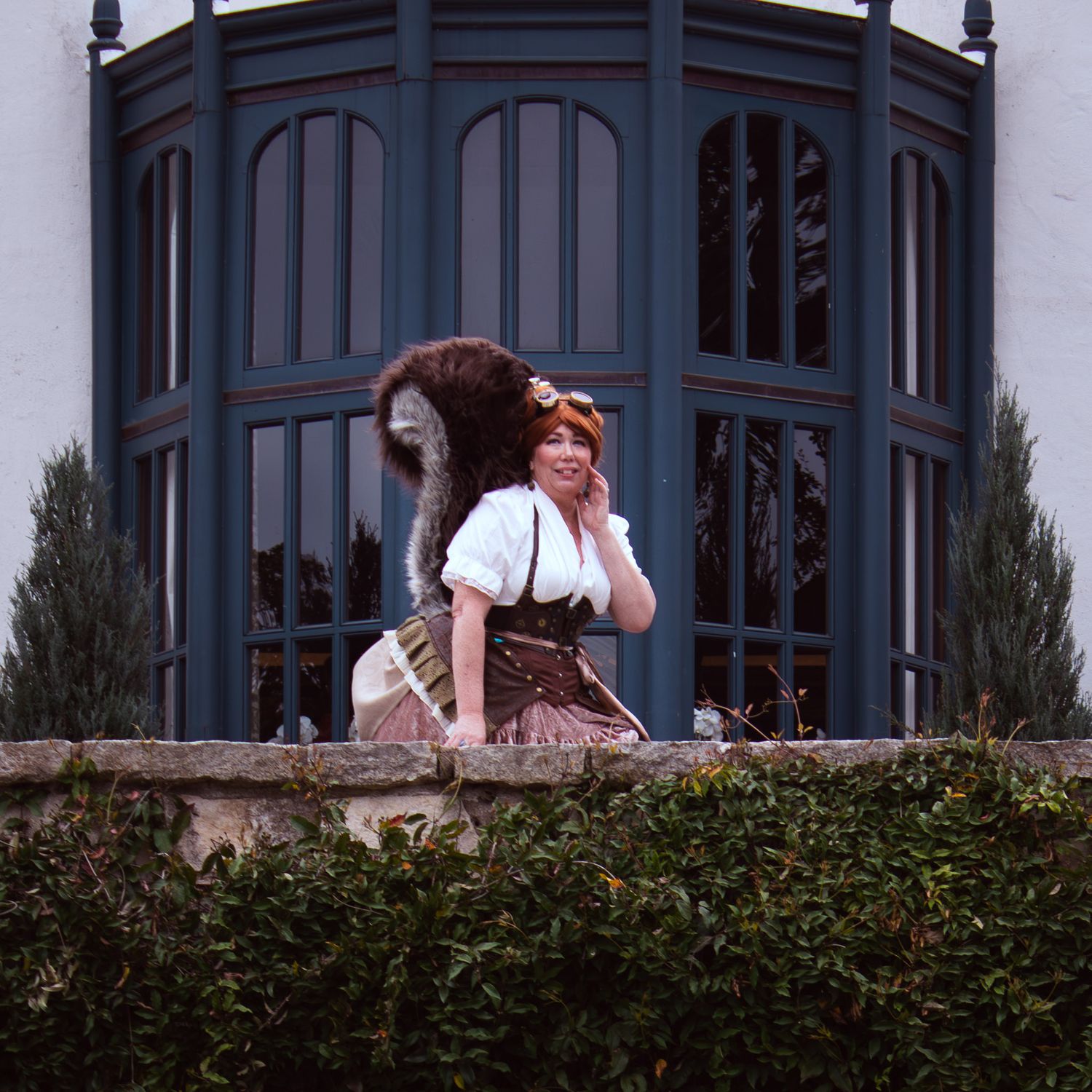 Woman in Renaissance costume with squirrel tail poses on stone ledge before ornate blue bay window.