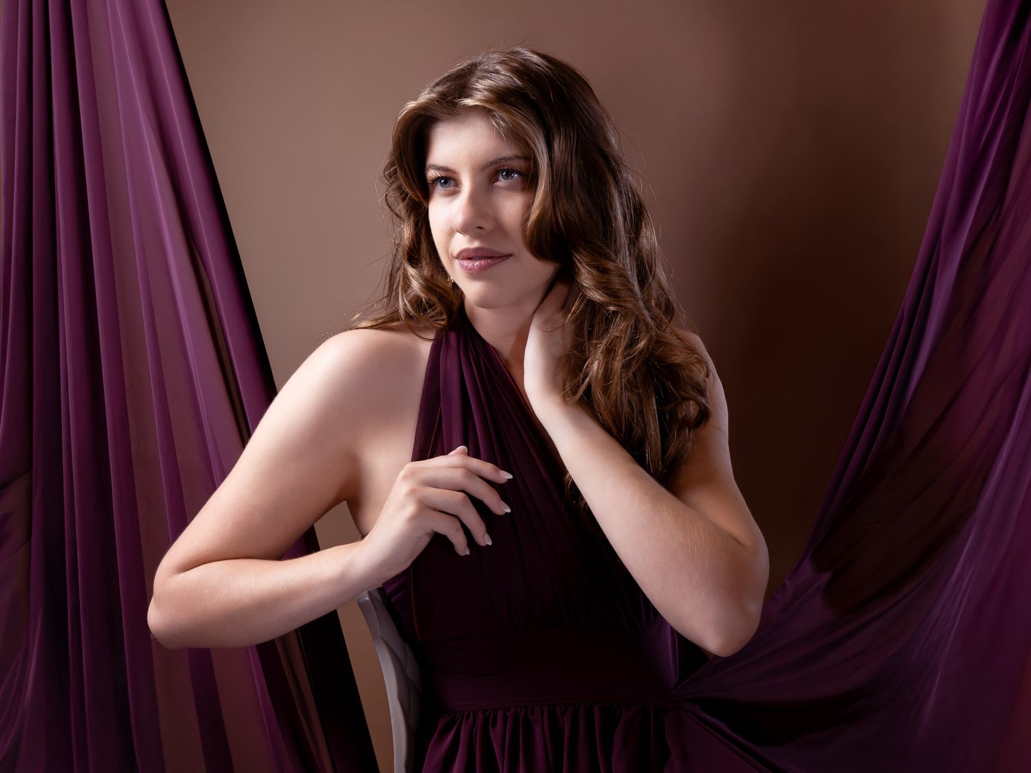 Woman in elegant purple halter dress poses gracefully against a mauve backdrop with flowing fabric drapes.