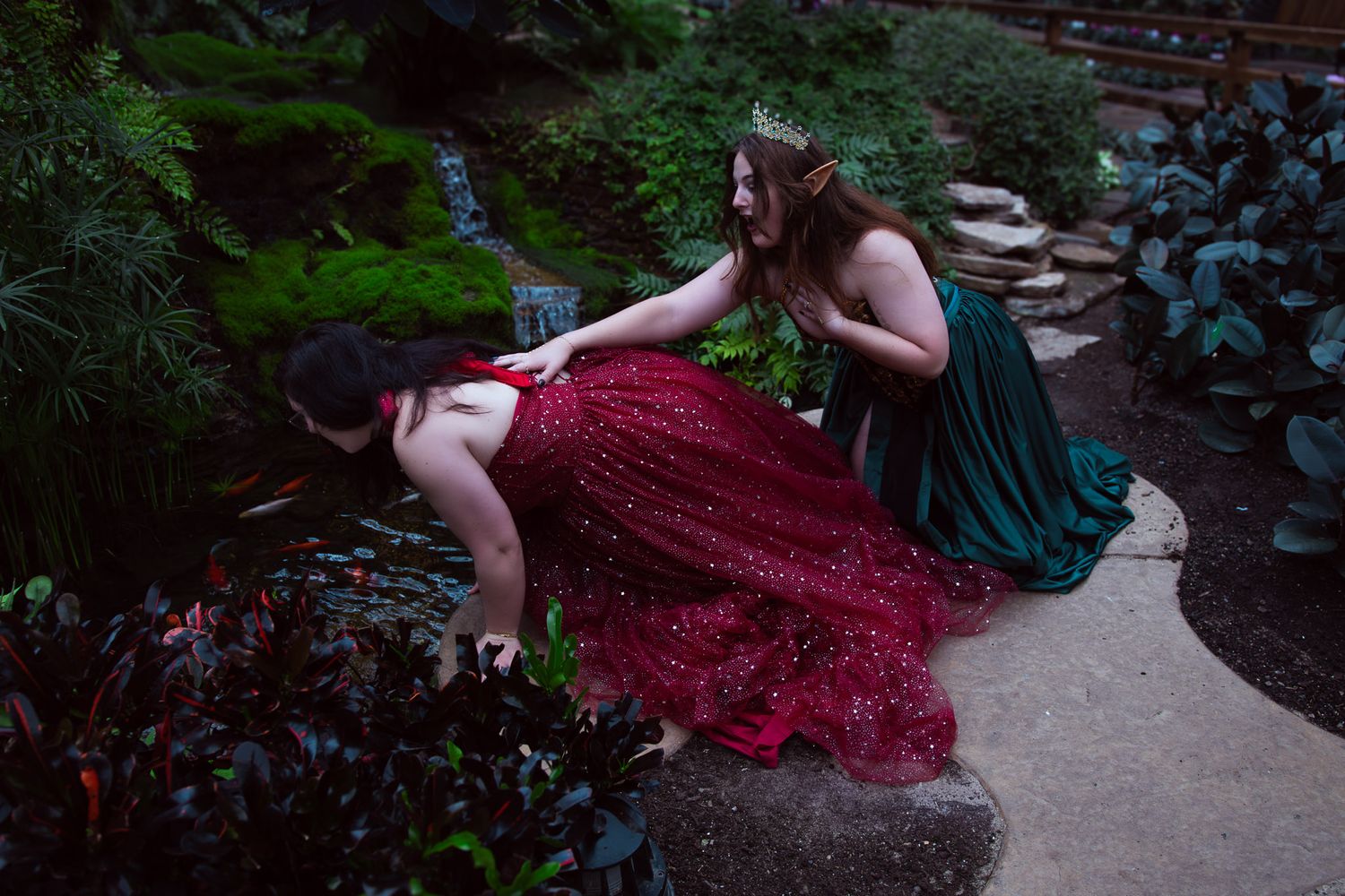 Two women in elegant red and green gowns pose in a lush garden setting during a fantasy-themed photo shoot.