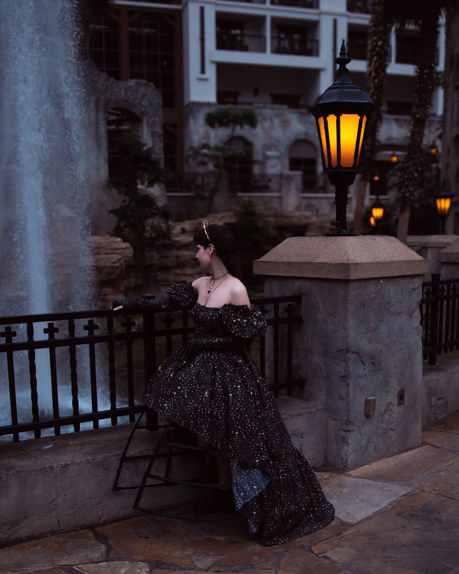 Woman in black off-shoulder gown poses by iron fence near glowing lantern at elegant outdoor venue.