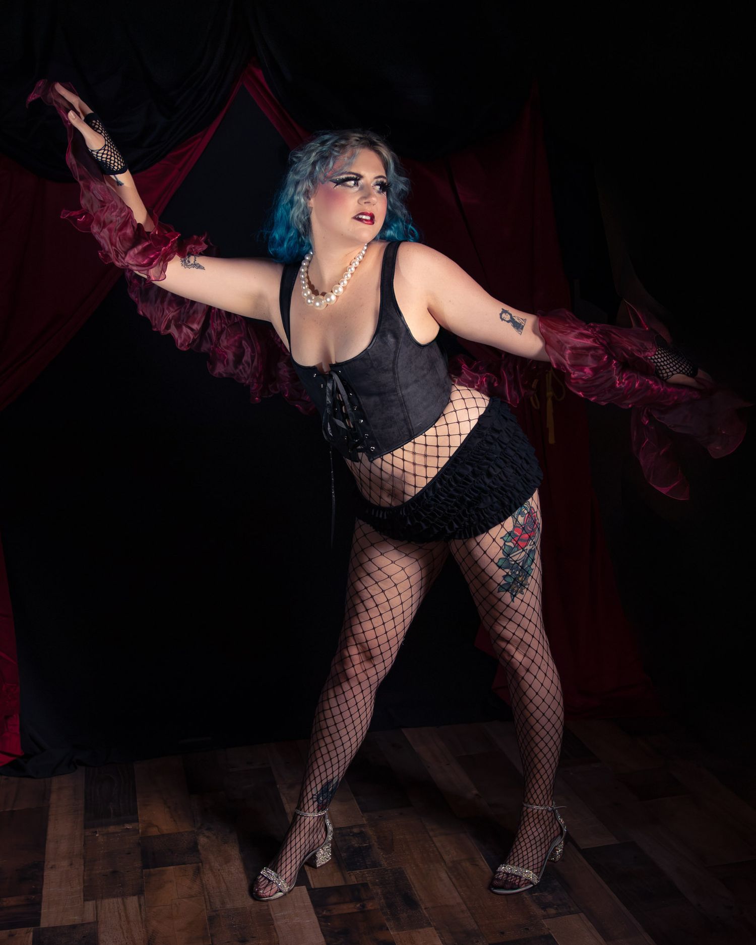 Blue-haired performer in black corset, fishnet stockings, and lace skirt poses dramatically on stage with red curtains.