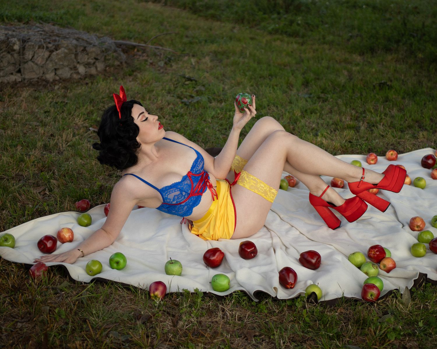 Woman in a Snow White-inspired costume with a red bow reclines on a white blanket surrounded by red and green apples on grass.