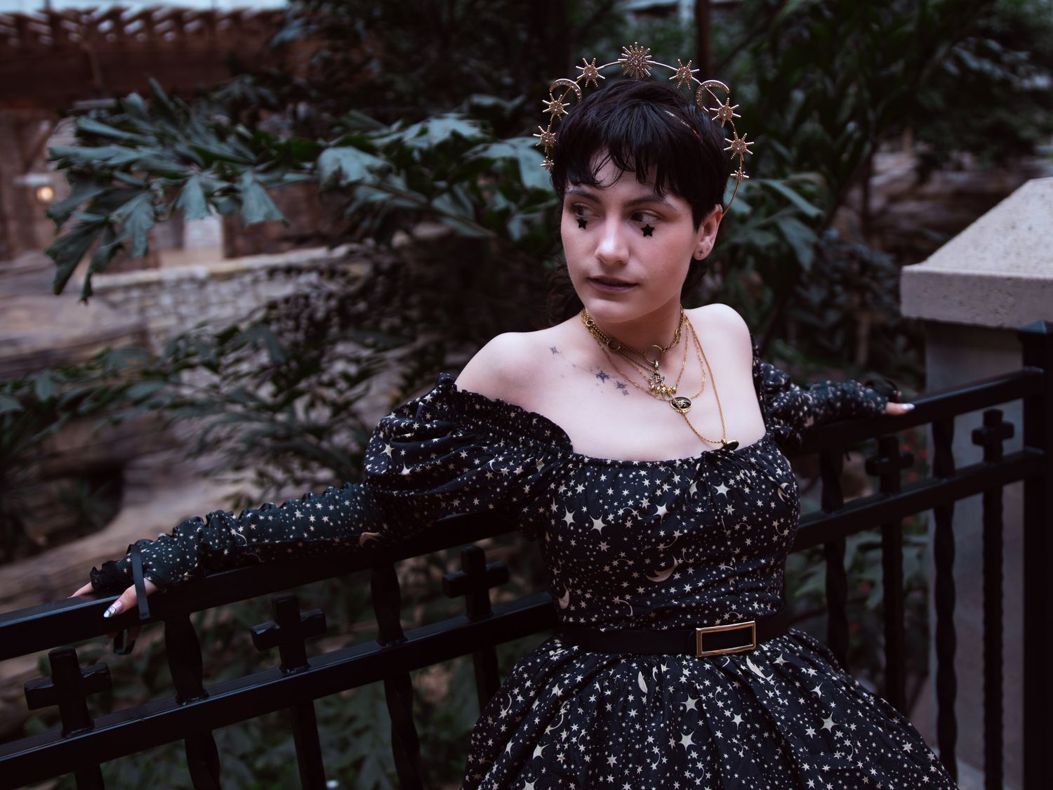 Woman in black star-print off-shoulder gown with gold crown poses elegantly by a railing outdoors.